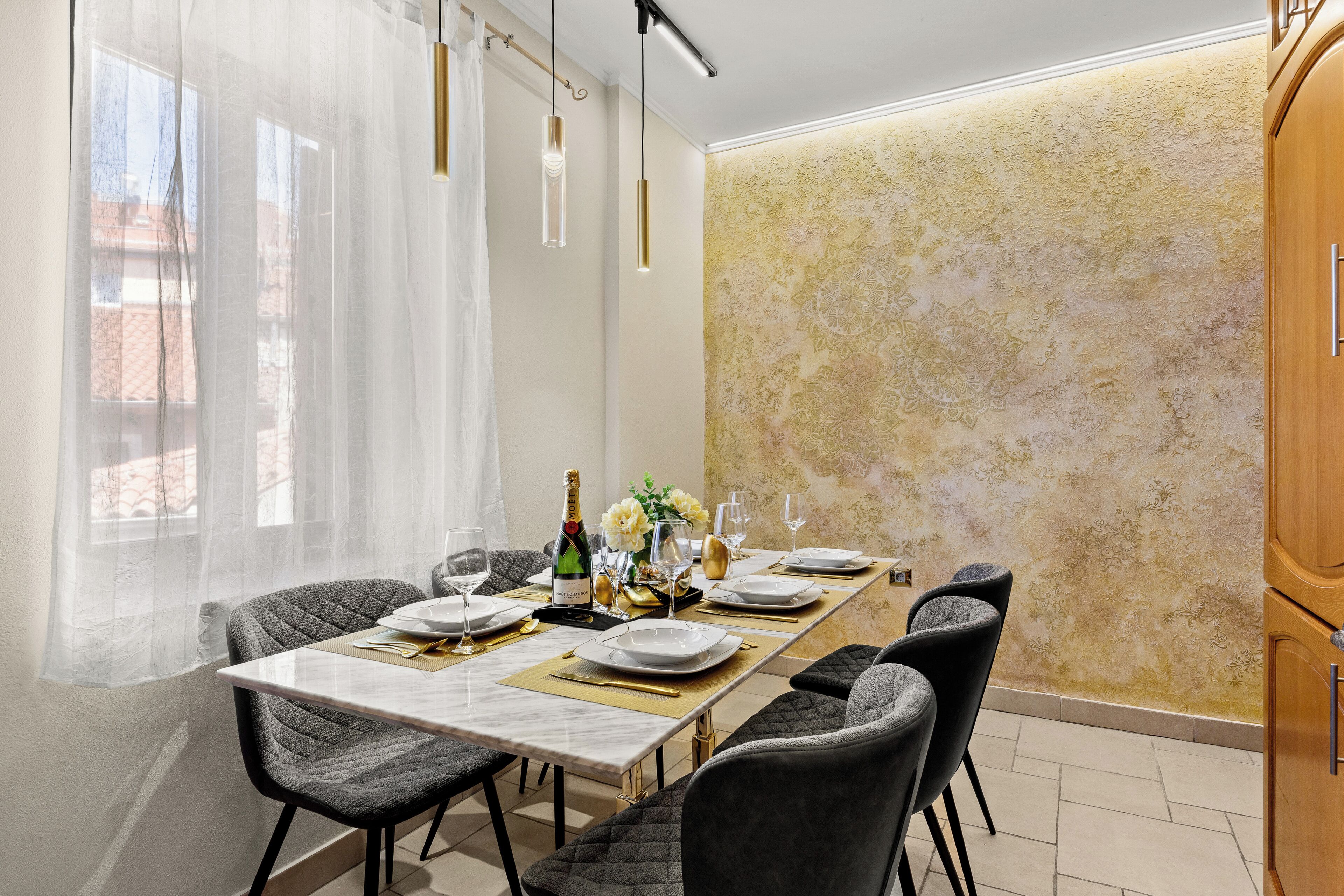A dining area with a long table set for four, featuring a bottle of champagne, glasses, and a vase of flowers.