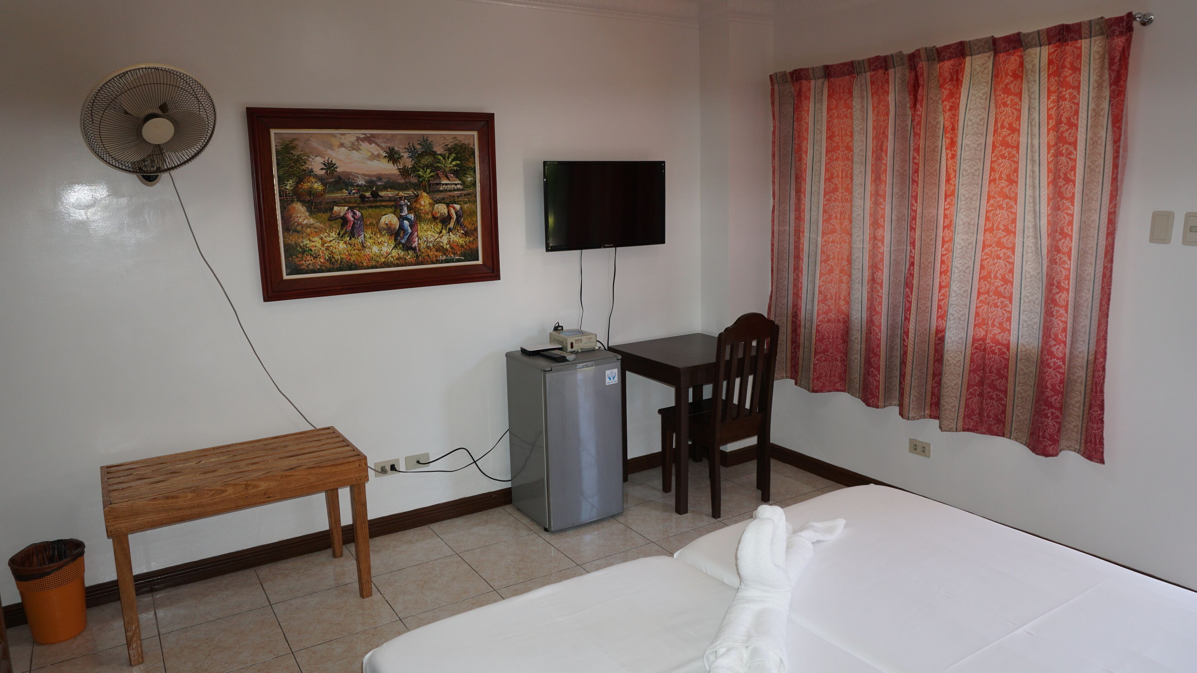 Habitación de hotel con una cama, un escritorio con computadora, una silla, un refrigerador pequeño, un cesto de basura, un ventilador y un cuadro en la pared.