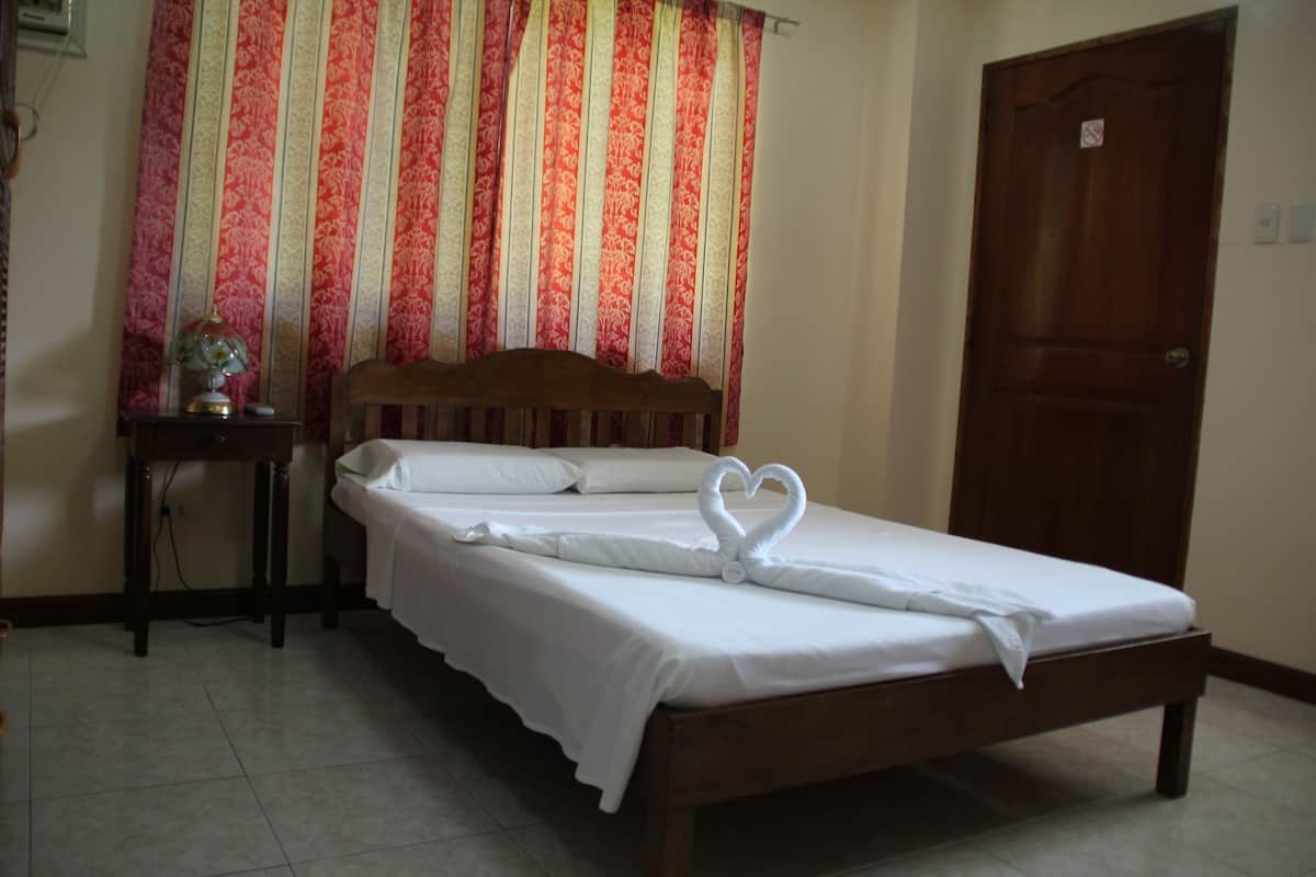 A neatly made bed with a heart-shaped towel on the bedspread, a wooden headboard, a nightstand with a lamp, and a door with a sign.