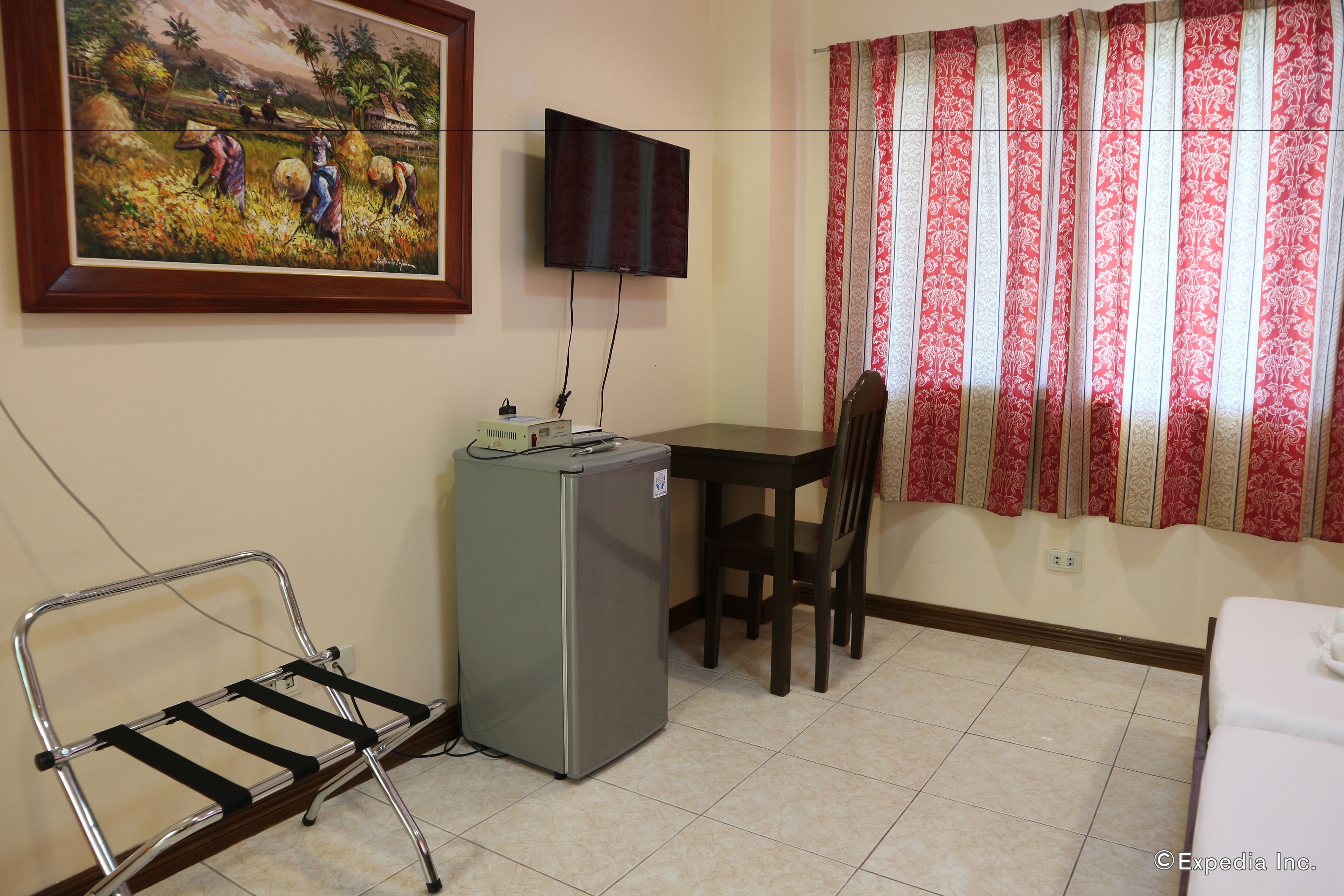 Une pièce comprenant un lit, un bureau, une chaise, un téléviseur fixé au mur et un tableau représentant des personnes travaillant dans un champ.