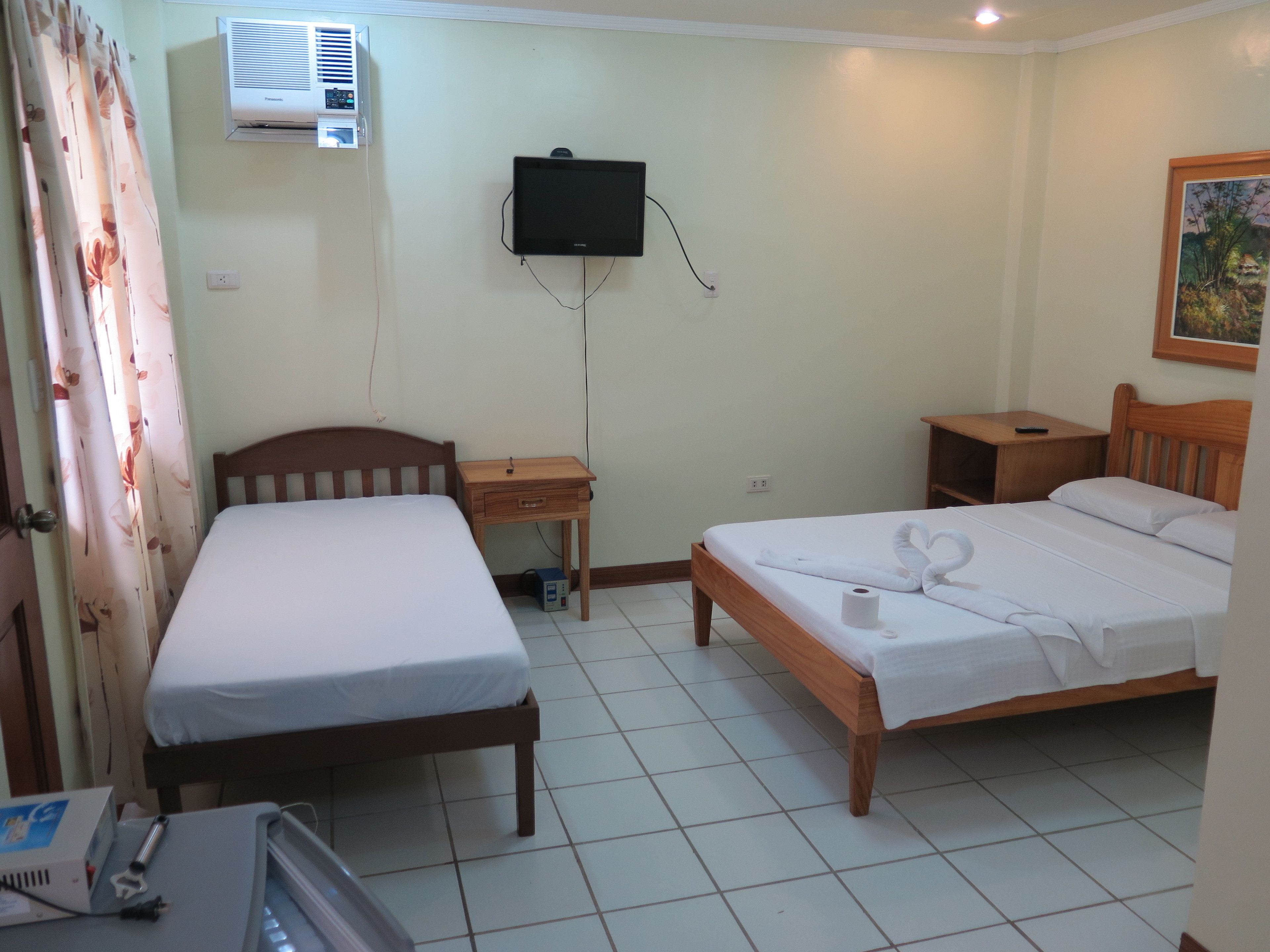 Habitación de hotel con dos camas individuales, una mesita de noche de madera, un televisor montado en la pared y un equipo de aire acondicionado.