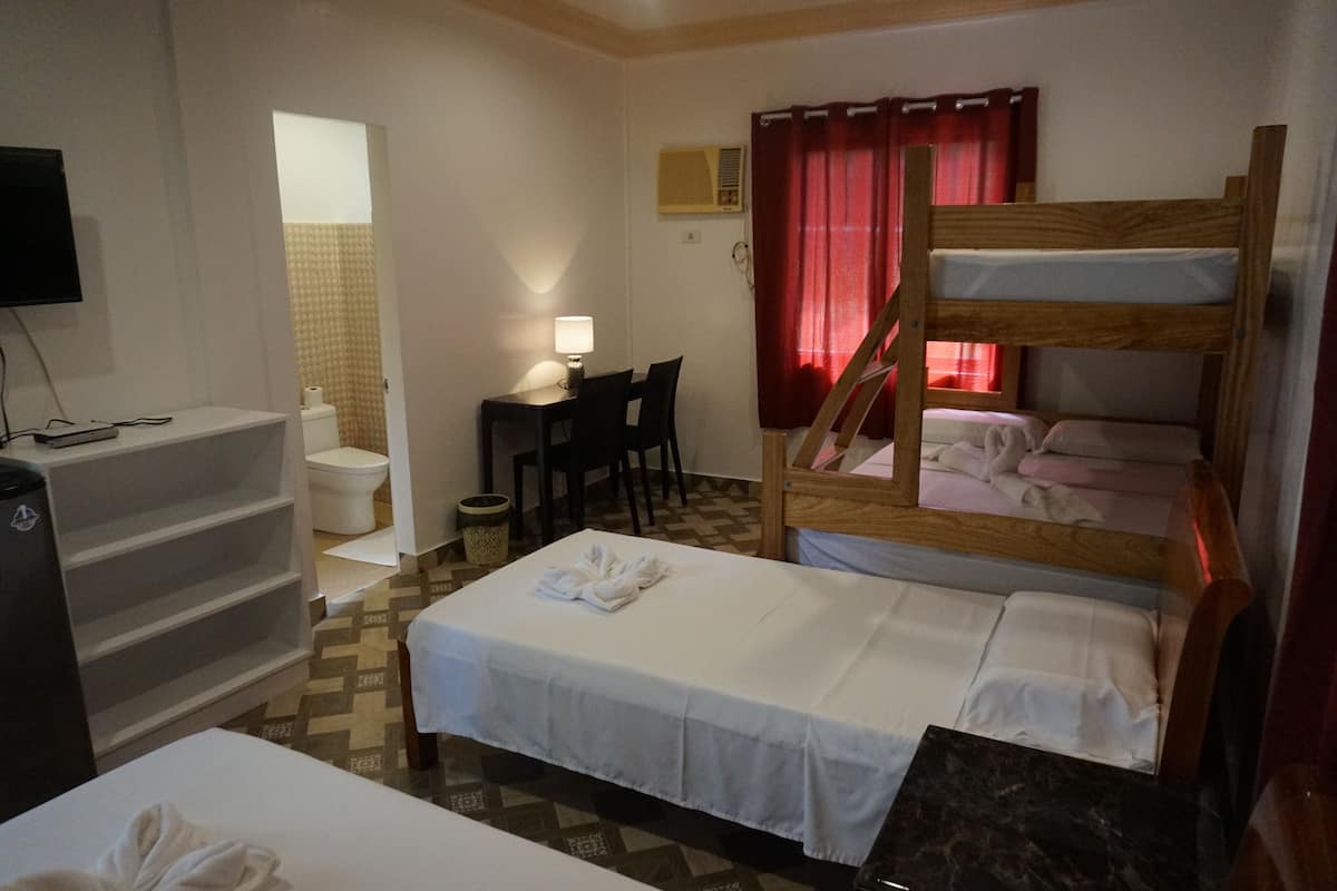 A hotel room with a bunk bed, a desk with a chair, a small table, a TV, and a bathroom visible through an open door.