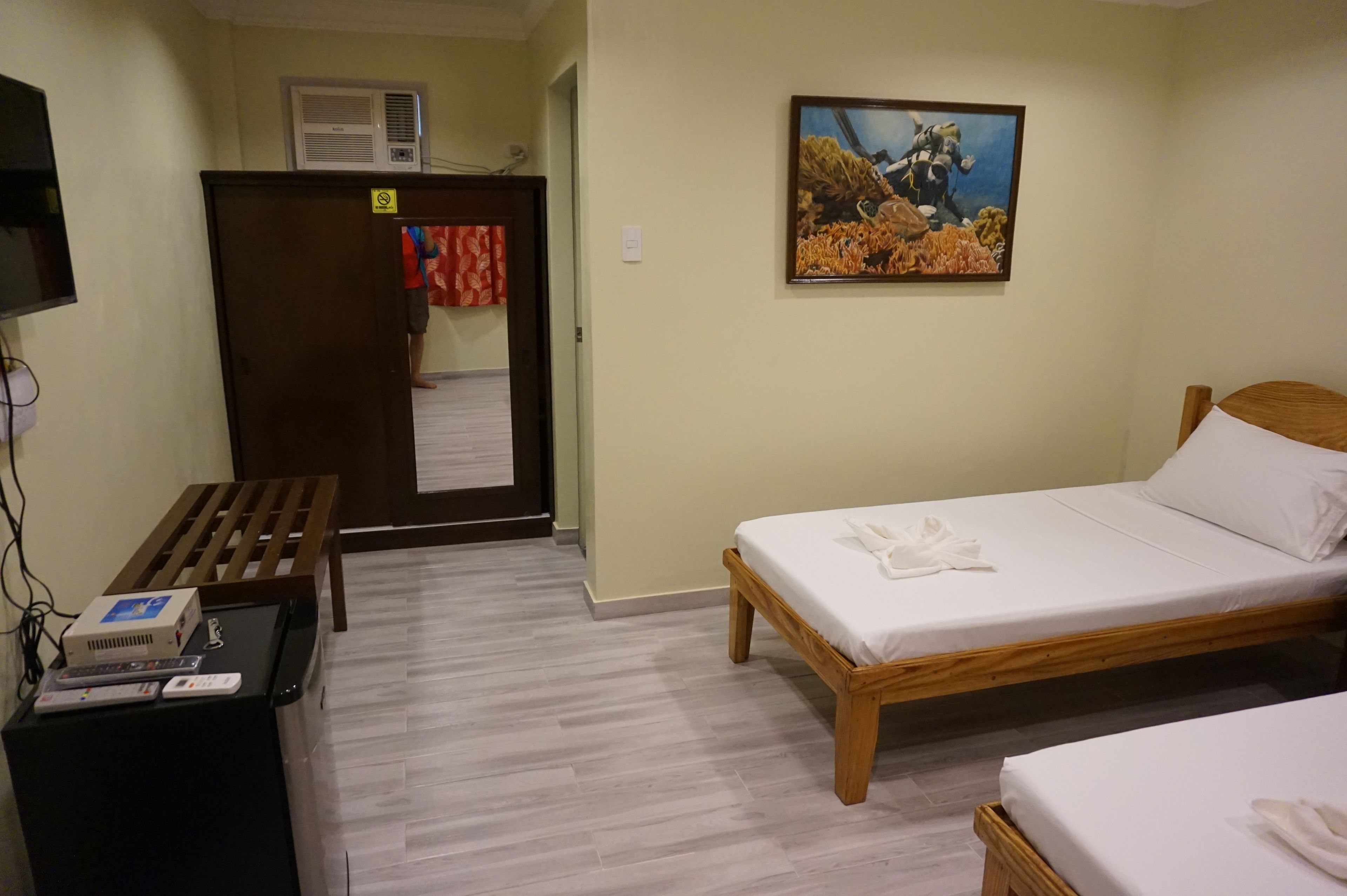 A hotel room with a bed, a desk with a computer and a projector, a wardrobe, and a painting on the wall.