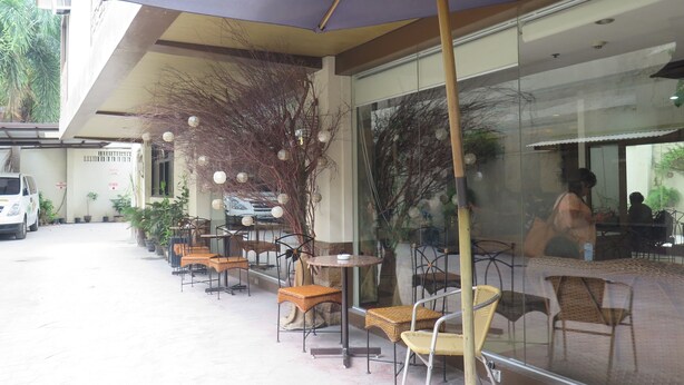 Un área de café al aire libre con mesas y sillas, un paraguas grande y un muro de vidrio que refleja el interior.