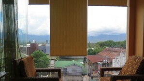Una habitación con dos sillas de mimbre, un ventanal con vistas a edificios y montañas, y una cortina.