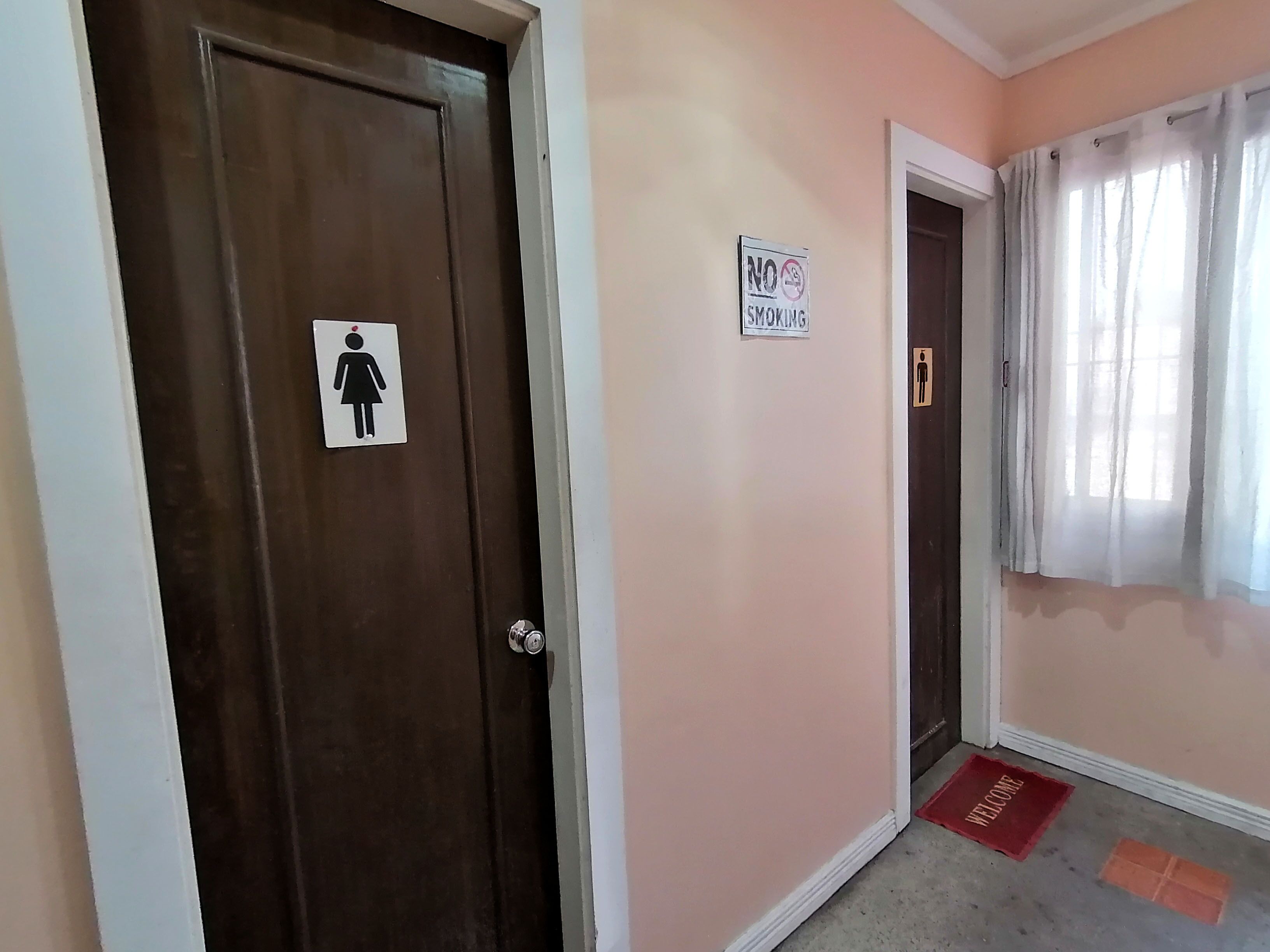 Un couloir avec une porte marron, un panneau « Interdiction de fumer », un symbole indiquant les toilettes pour femmes et un paillasson avec l’inscription « Bienvenue ».