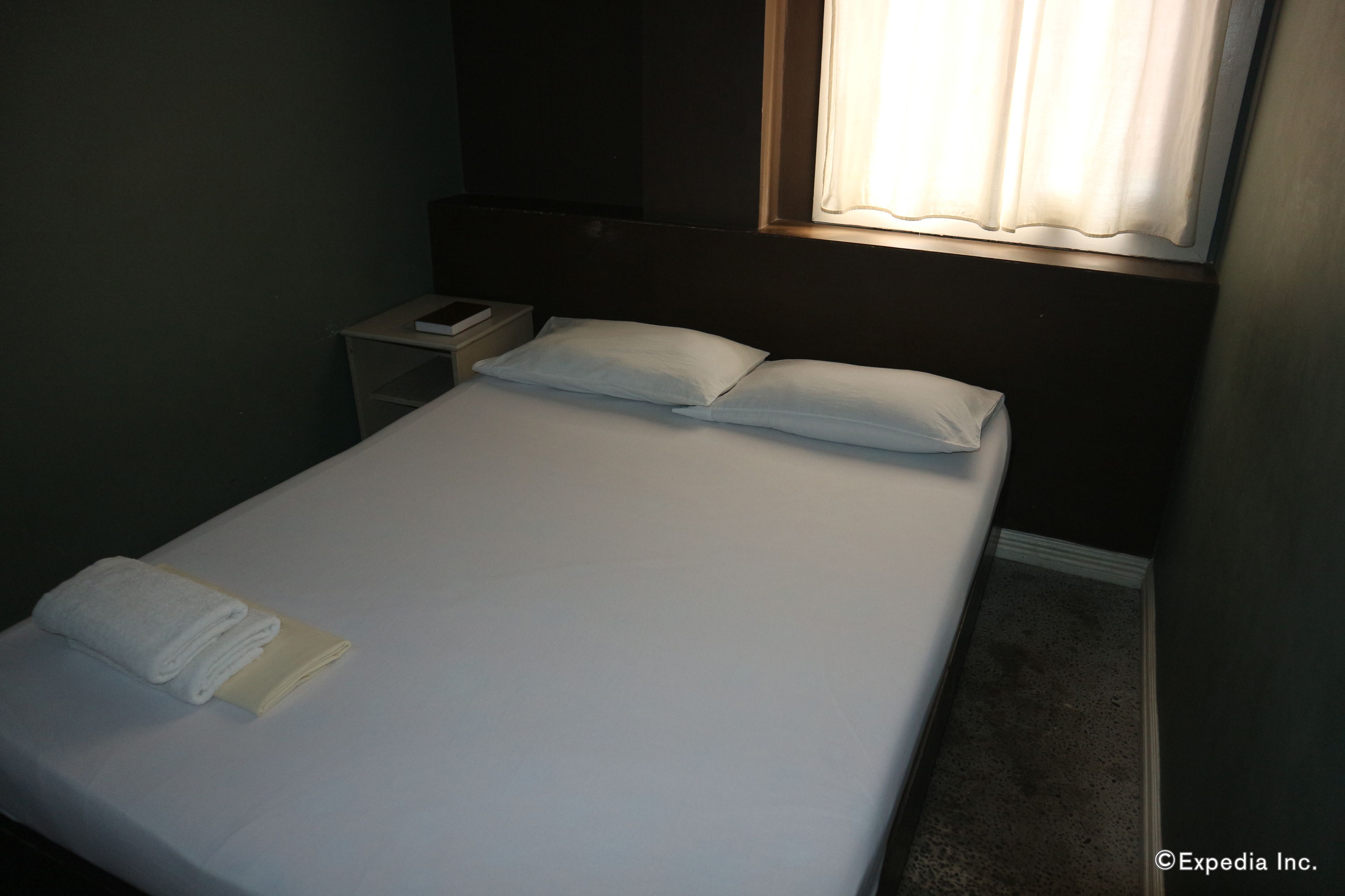 Una cama bien hecha con sábanas y almohadones blancos, una mesita de noche con un libro y un teléfono, y una ventana con cortinas.