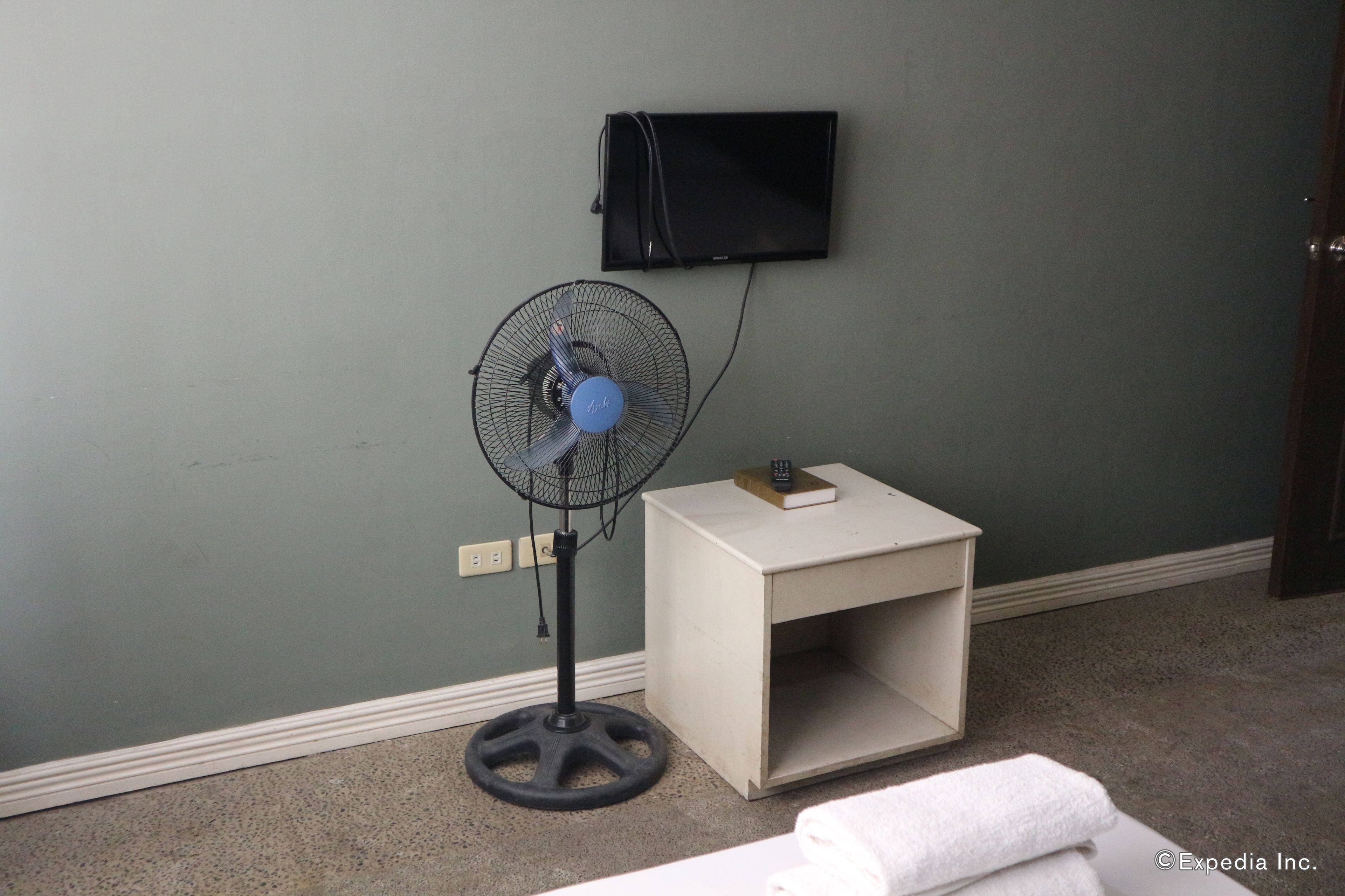 Une pièce avec un ventilateur sur pied, une petite table de chevet blanche et un téléviseur fixé au mur.