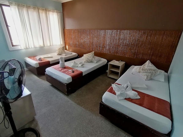 A hotel room with three single beds, a small nightstand, a fan, and a window with curtains.