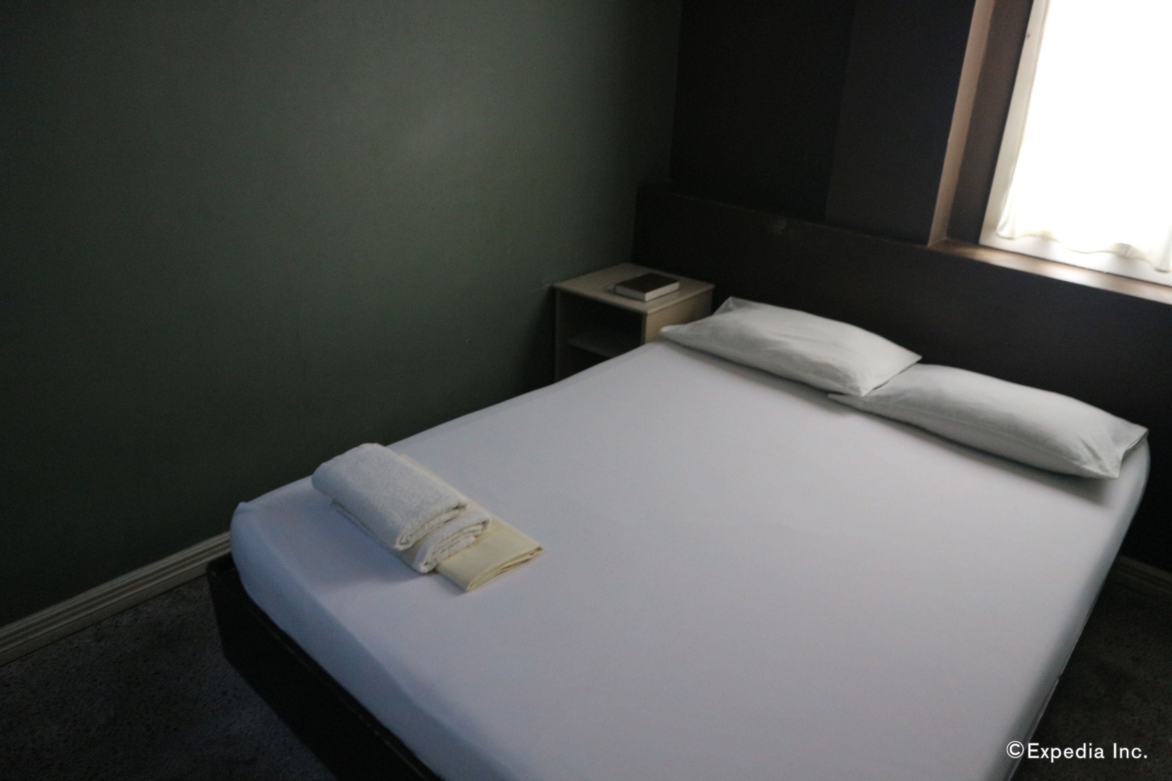Un letto rifatto con lenzuola e cuscini bianchi, un comodino con un libro e un telefono, e una finestra con tende.