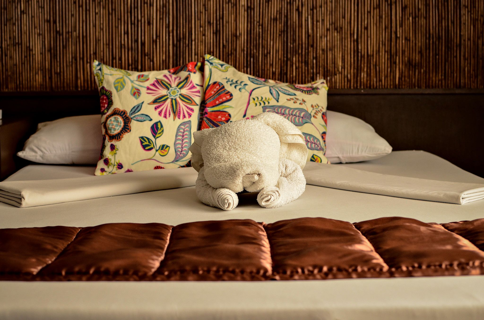 A neatly made bed with a plush white teddy bear, colorful floral pillows, and a brown leather headboard.