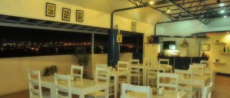 Un restaurante con mesas y sillas blancas, vistas a la ciudad de noche y un techo alto con vigas a la vista.