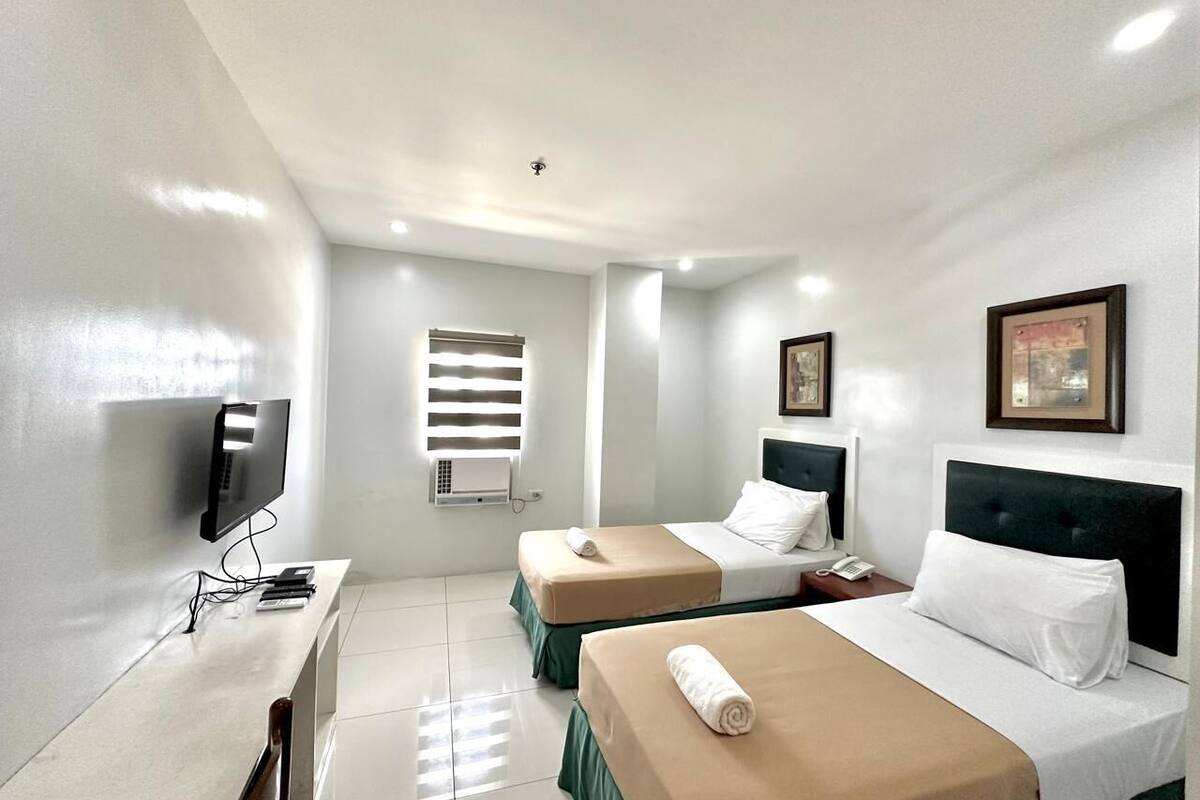 A hotel room with two beds, a desk with a computer, a TV, and a window with blinds.