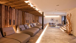 Uma área de spa com várias cadeiras de massagem dispostas ao longo de uma parede de madeira, uma área de estar com mesa e cadeiras, e uma escultura de madeira decorativa à direita.