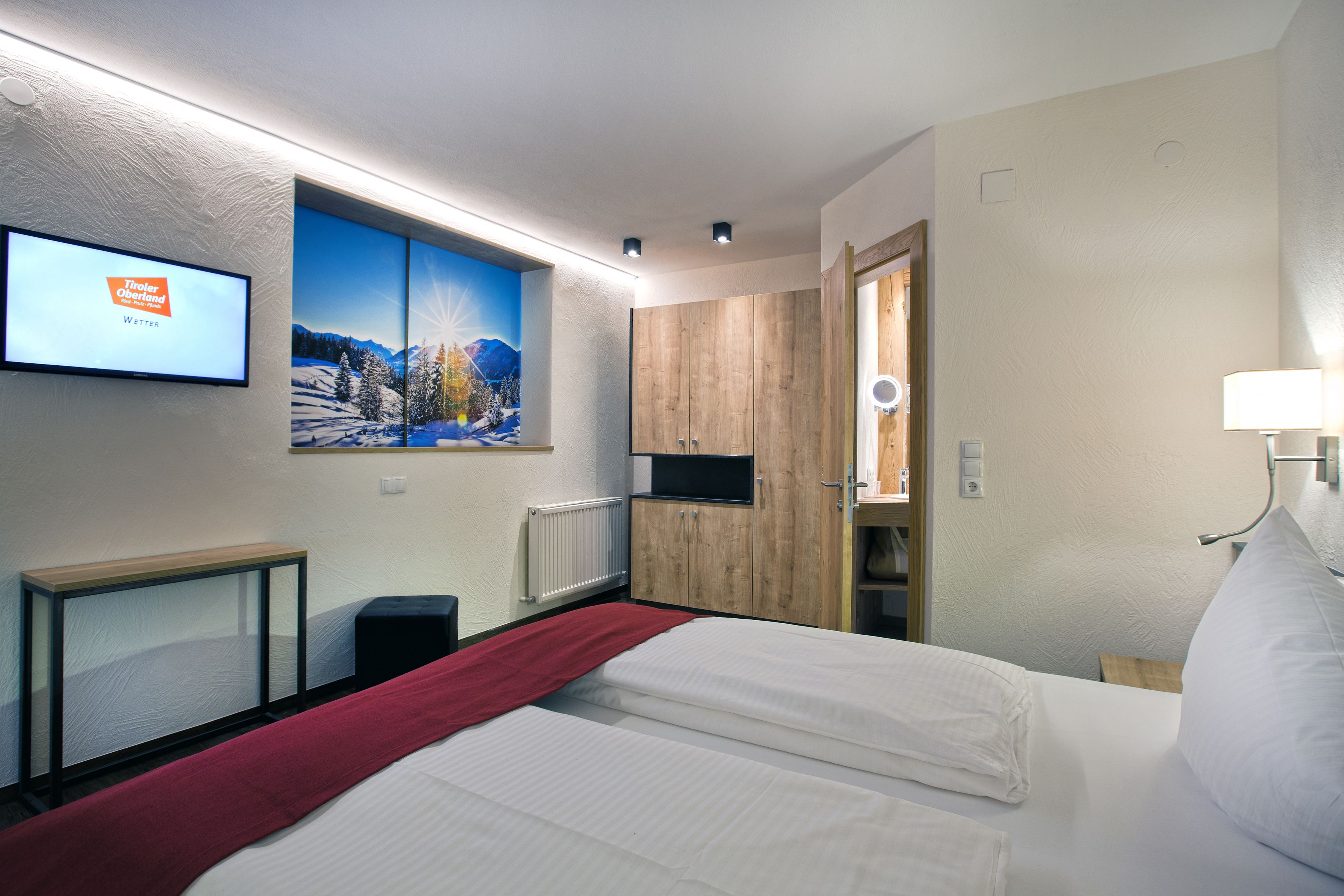 Ein Hotelzimmer mit einem Bett, einem an der Wand befestigten Fernseher, einem Holzschrank und einer roten Decke auf dem Bett.
