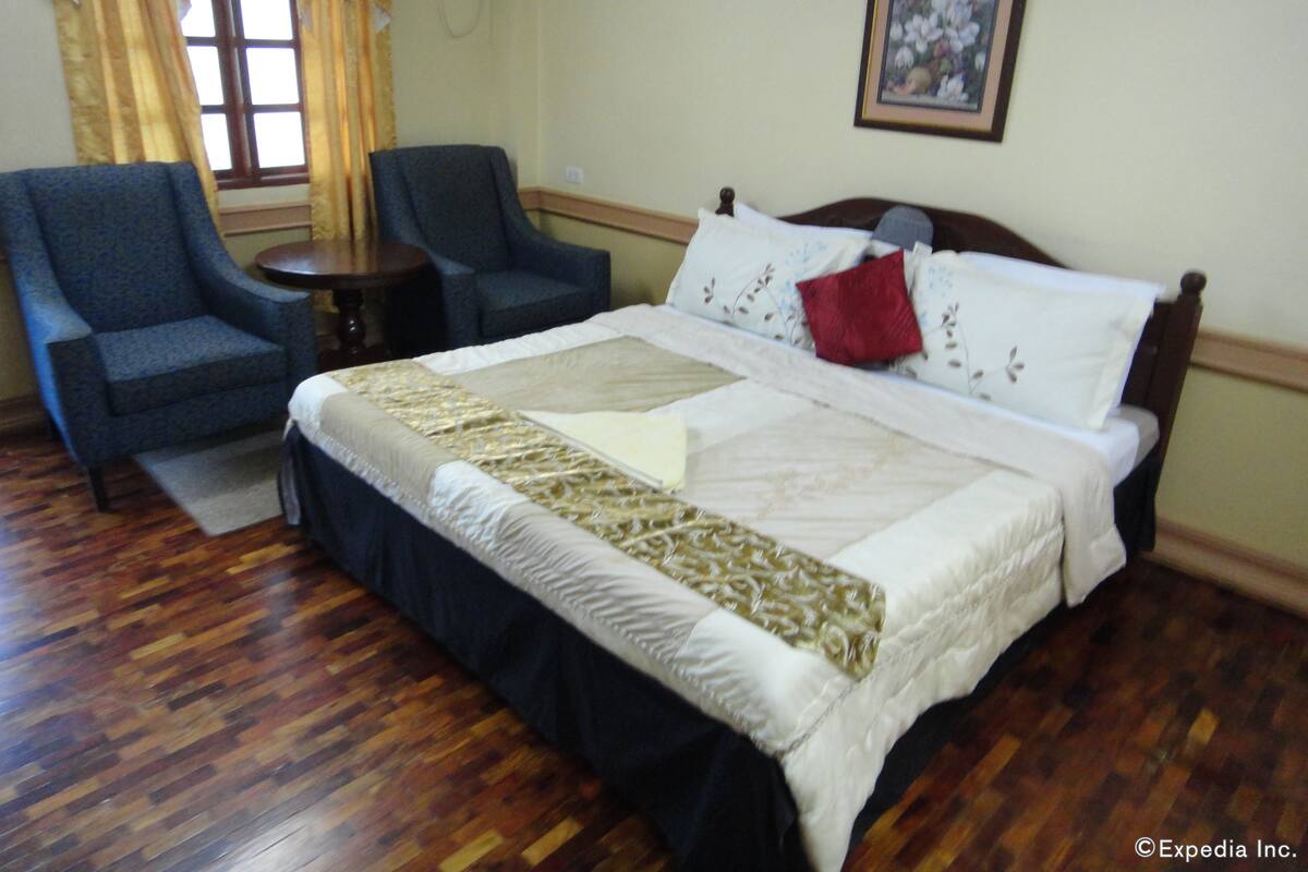 Habitación de hotel con una cama grande, dos sillones, una mesita redonda y un ventanal con cortinas.