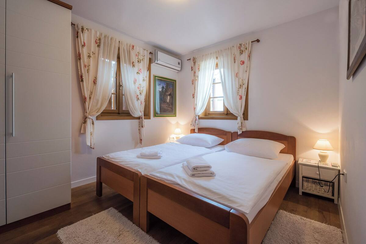 Ein Schlafzimmer mit einem Doppelbett, einem Holz-Kopfteil, zwei Nachttischlampen, einem kleinen Nachttisch, einer Wand-Klimaanlage und einem Fenster mit floralem Vorhang.