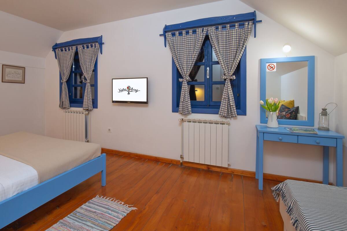 A bedroom with a bed, two windows with curtains, a radiator, a desk with a mirror, and a framed picture on the wall.