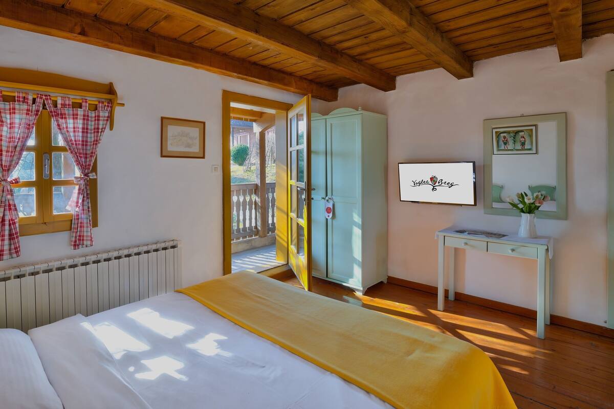 A bedroom with a bed, a radiator, a window with curtains, a wardrobe, a desk with a vase, and a framed picture.