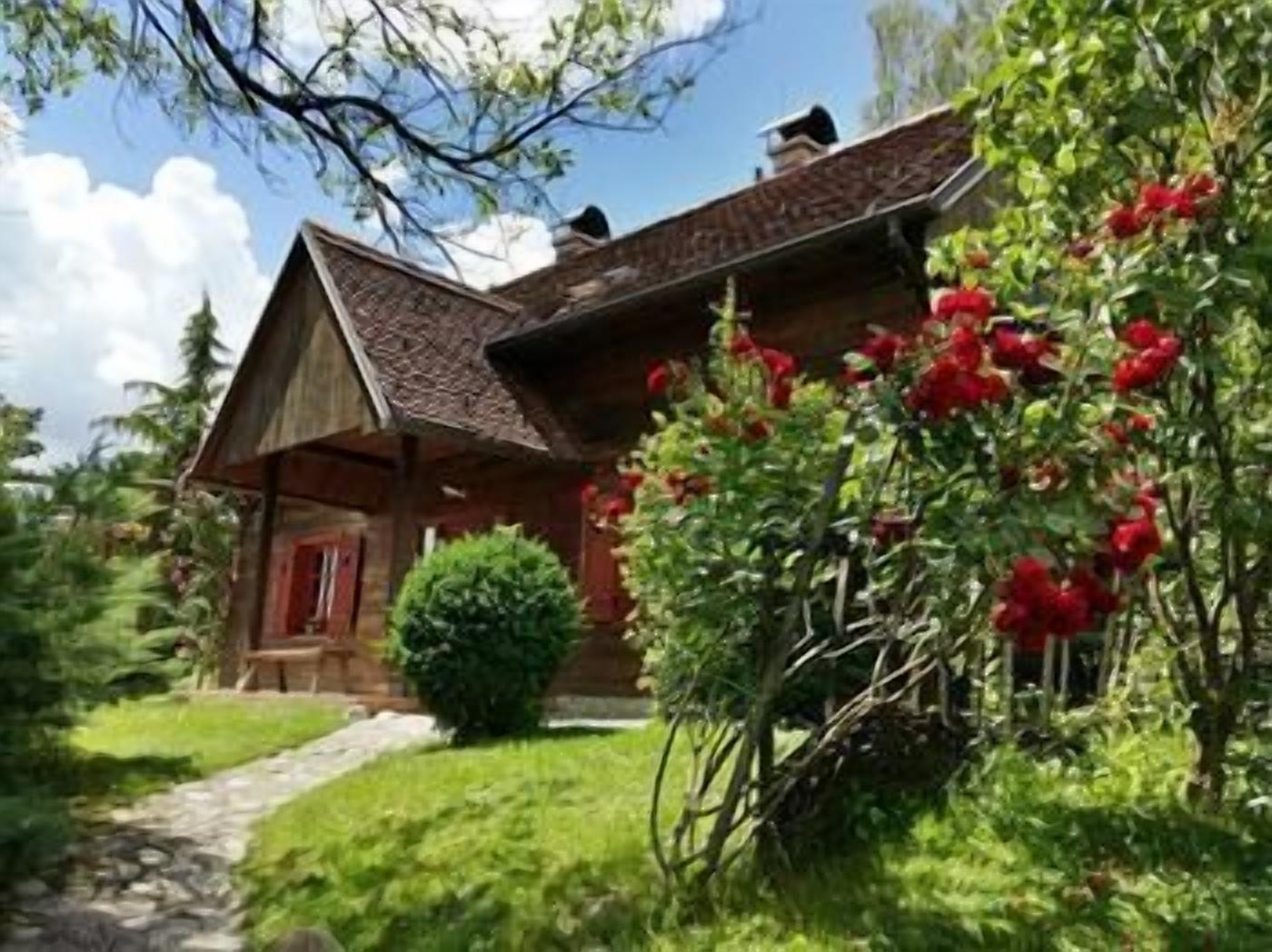 Une maison de campagne en bois, avec une porte rouge, entourée d’une végétation luxuriante et de fleurs rouges.