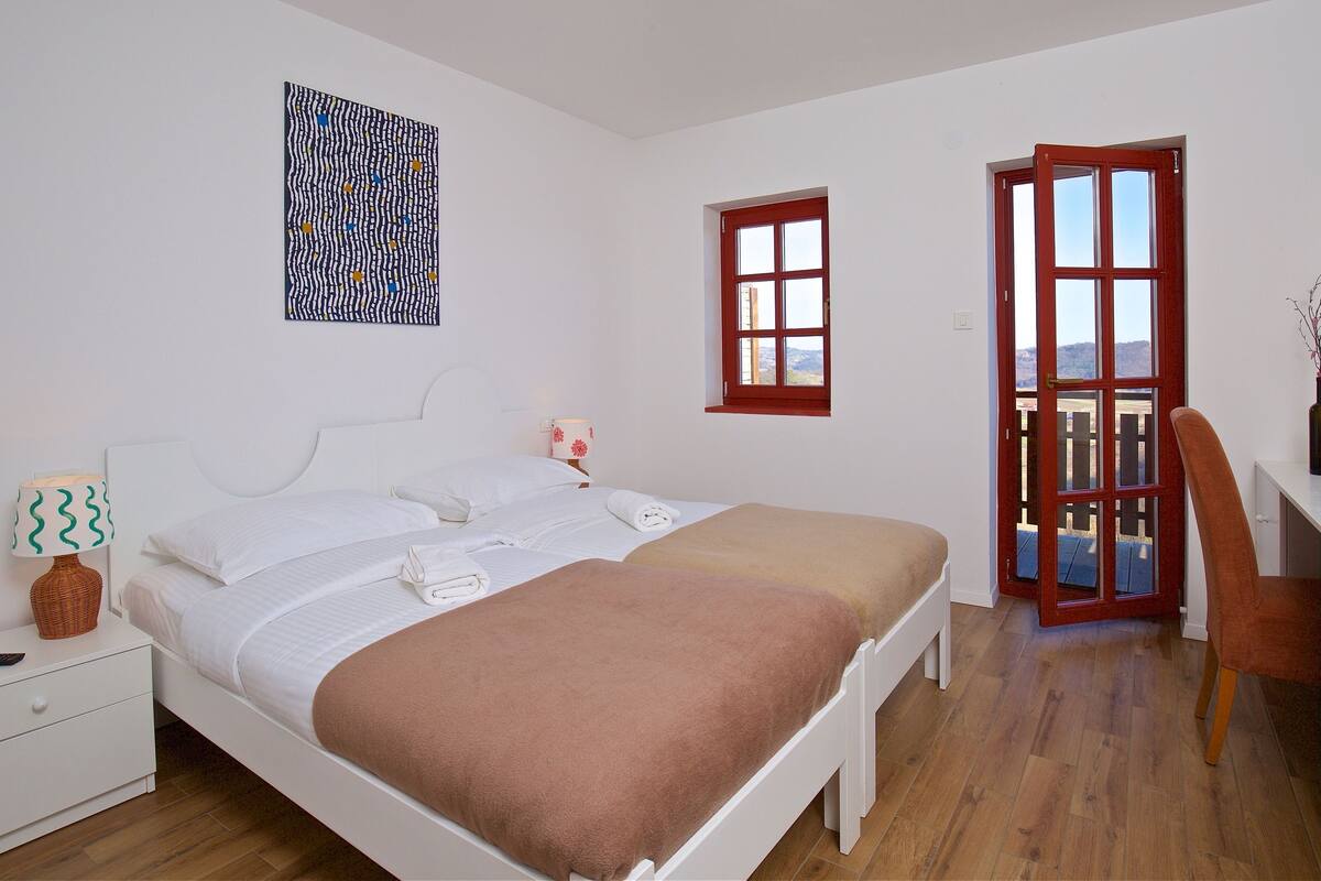 A bedroom with a bed, a desk, a chair, a nightstand, a lamp, a vase, and a window.