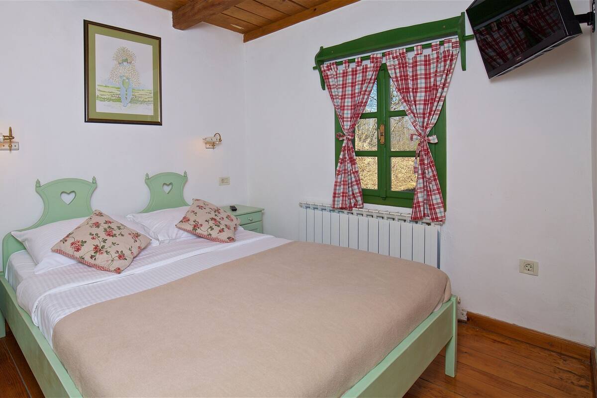 A bedroom with a bed, green headboard, bedside tables, a telephone, a framed picture, a window with curtains, and a radiator.