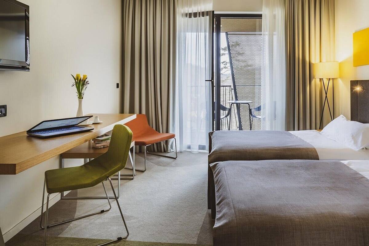 A hotel room with two beds, a desk with a laptop, a chair, a TV, and a balcony with a view.