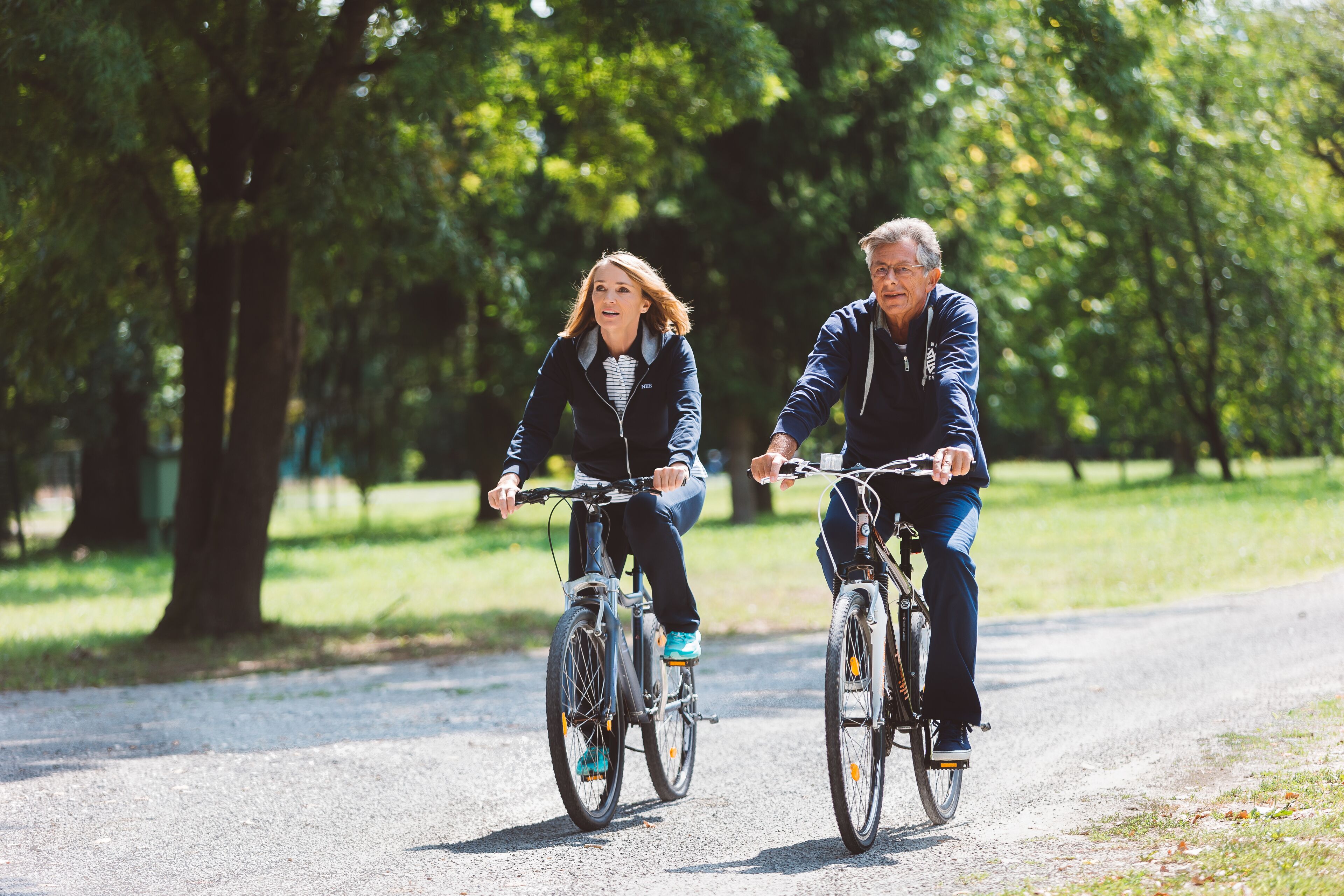 Deux personnes font du vélo sur un chemin de gravier dans un parc, au milieu des arbres.