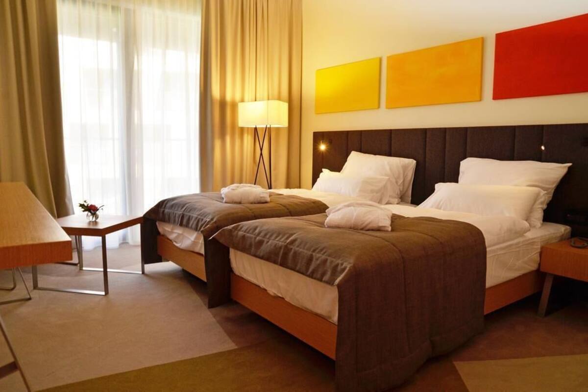 A hotel room with two beds, a bedside table, a lamp, and a view of the outside through a window with curtains.