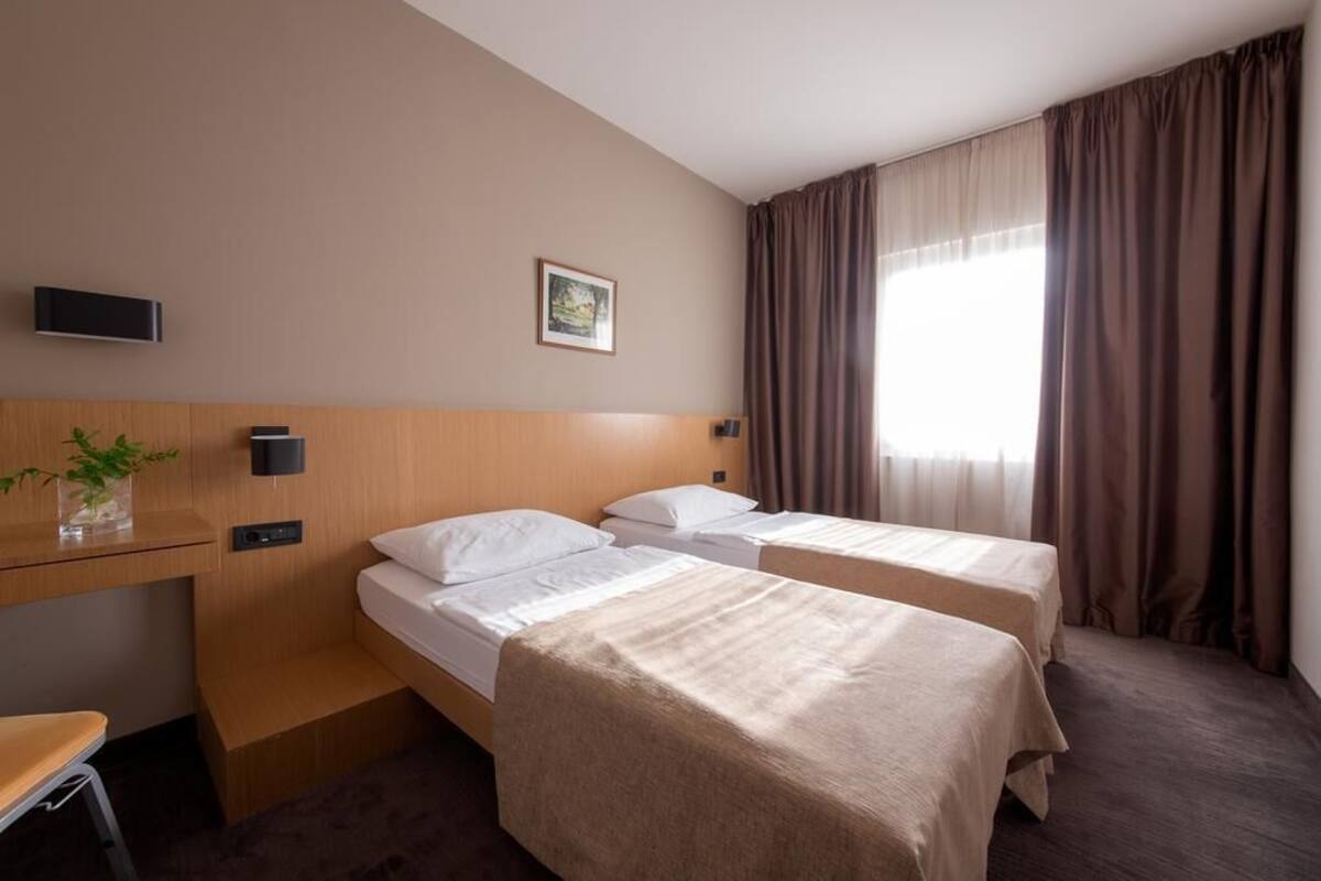 A hotel room with two single beds, a desk with a chair, a TV, and a window with curtains.