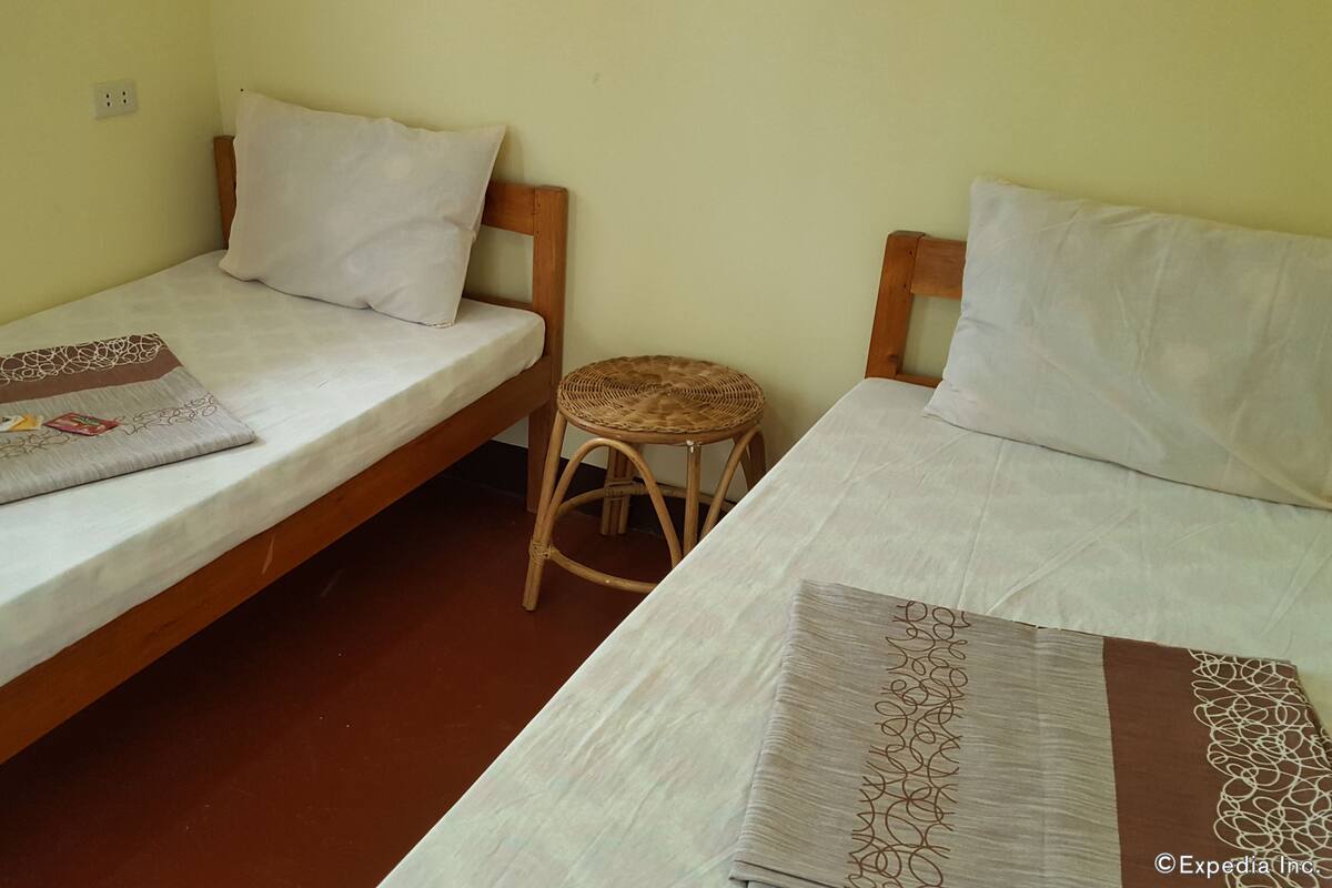 A small, simple room with two single beds, a small wooden stool, and a plain wall.