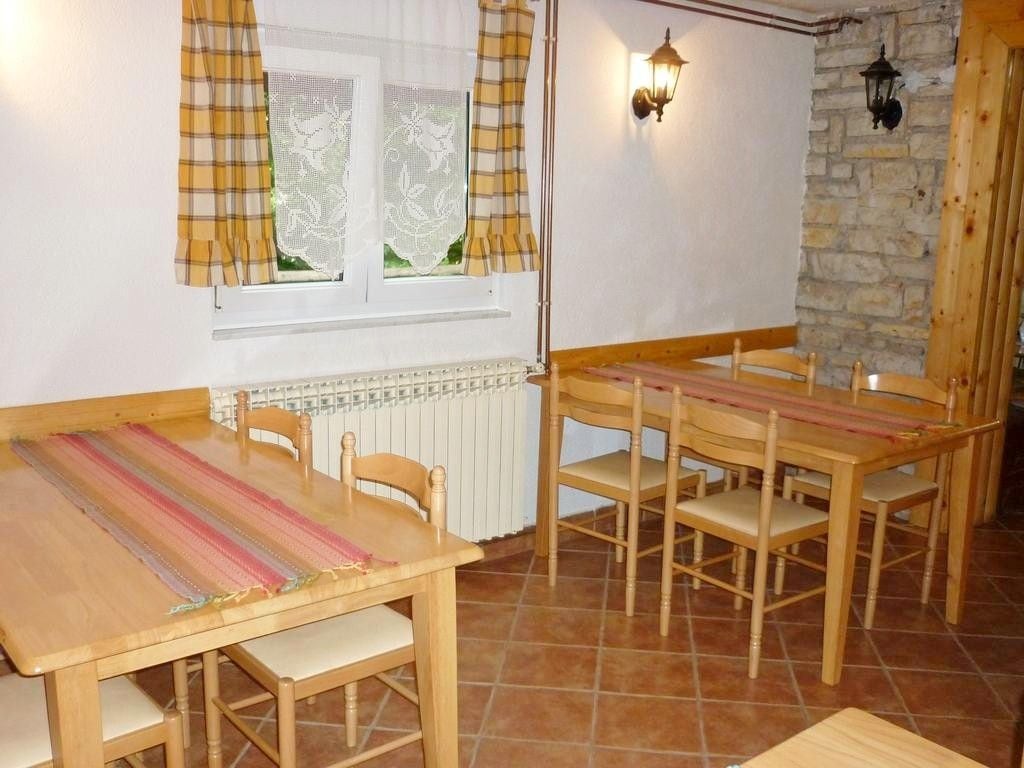 A dining area with wooden tables and chairs, a window with curtains, a wall-mounted light fixture, and a stone wall.