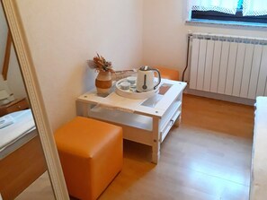 Una habitación pequeña y funcional con un radiador blanco, una mesa con un hervidor, tazas y un florero, y una silla naranja.