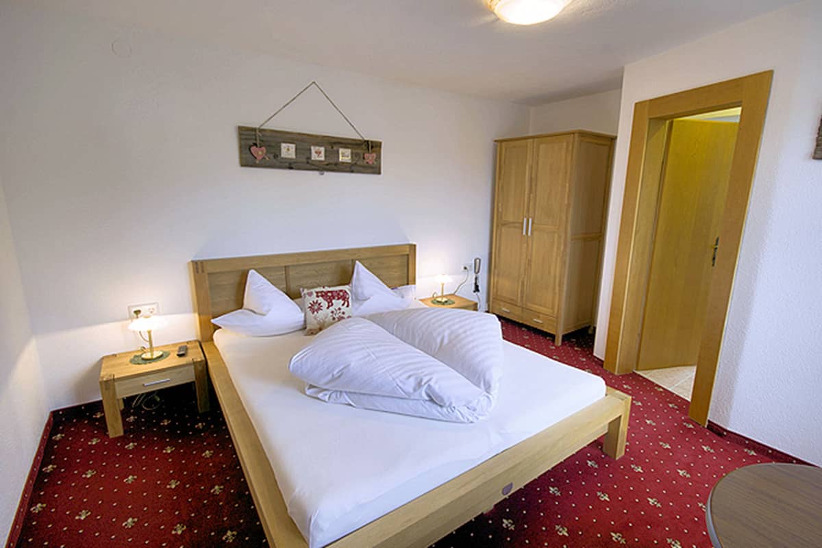 A neatly made bed with white linens, a wooden headboard, and a bedside table with a lamp. There is a wooden wardrobe and a door leading to another room.