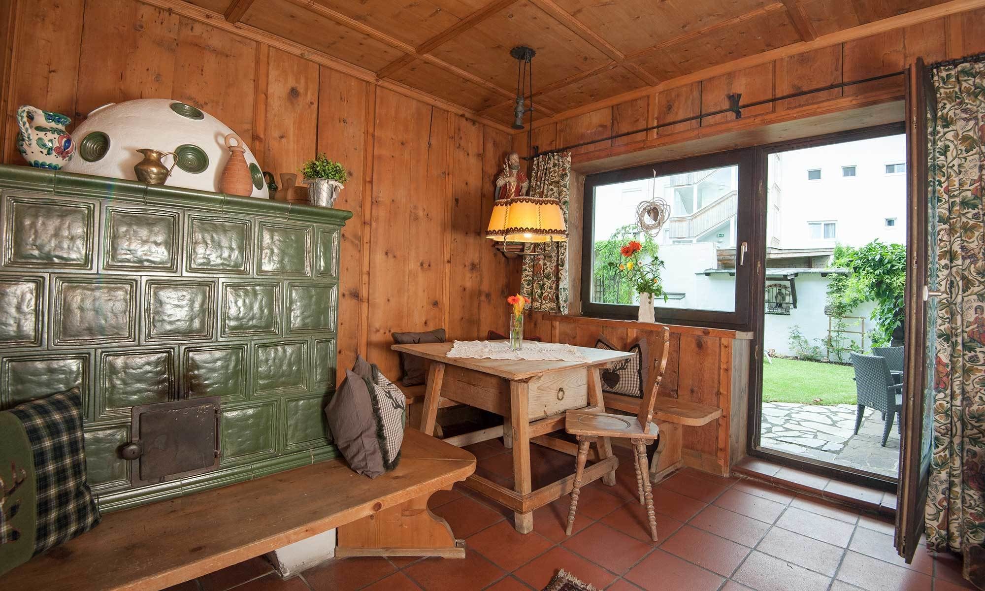 A cozy room with wooden walls, a green cabinet, a wooden table with chairs, a bench, and a window with a view of a garden and buildings.