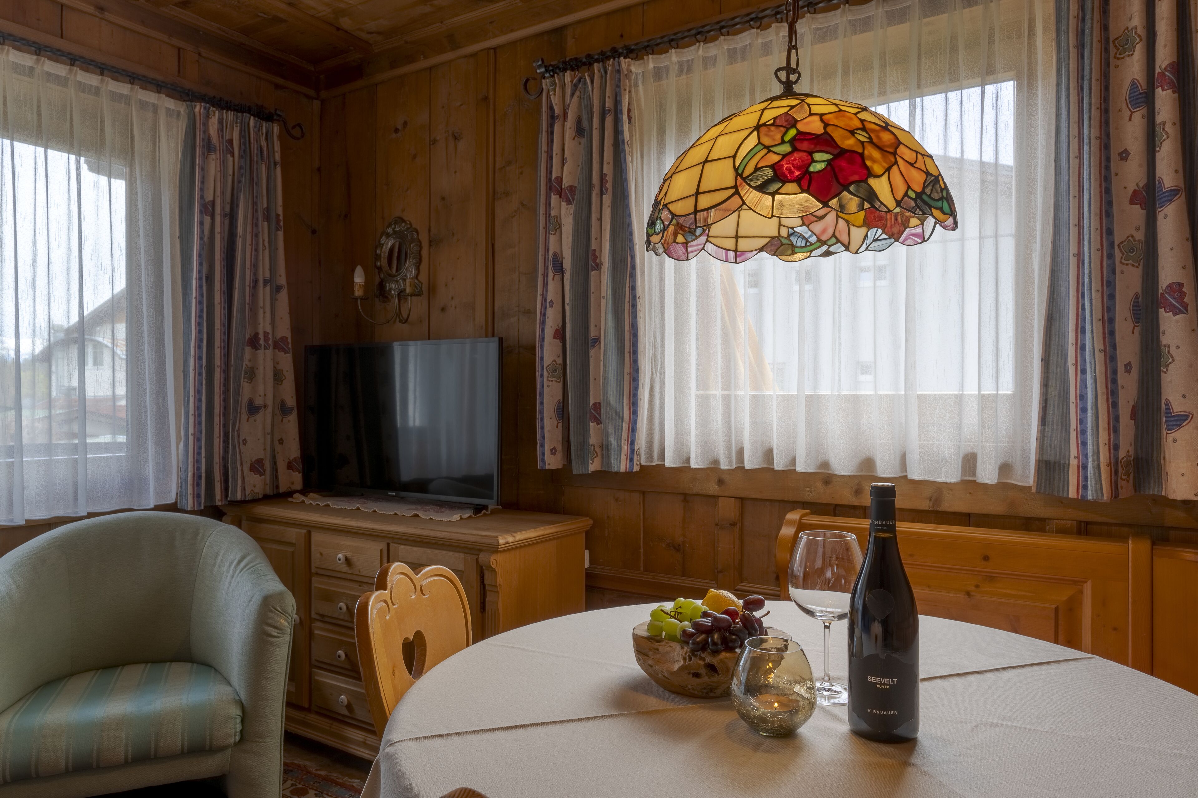 A cozy room with a wooden interior, a television, a dining table with a bowl of fruit and a bottle of wine, and a stained glass window.