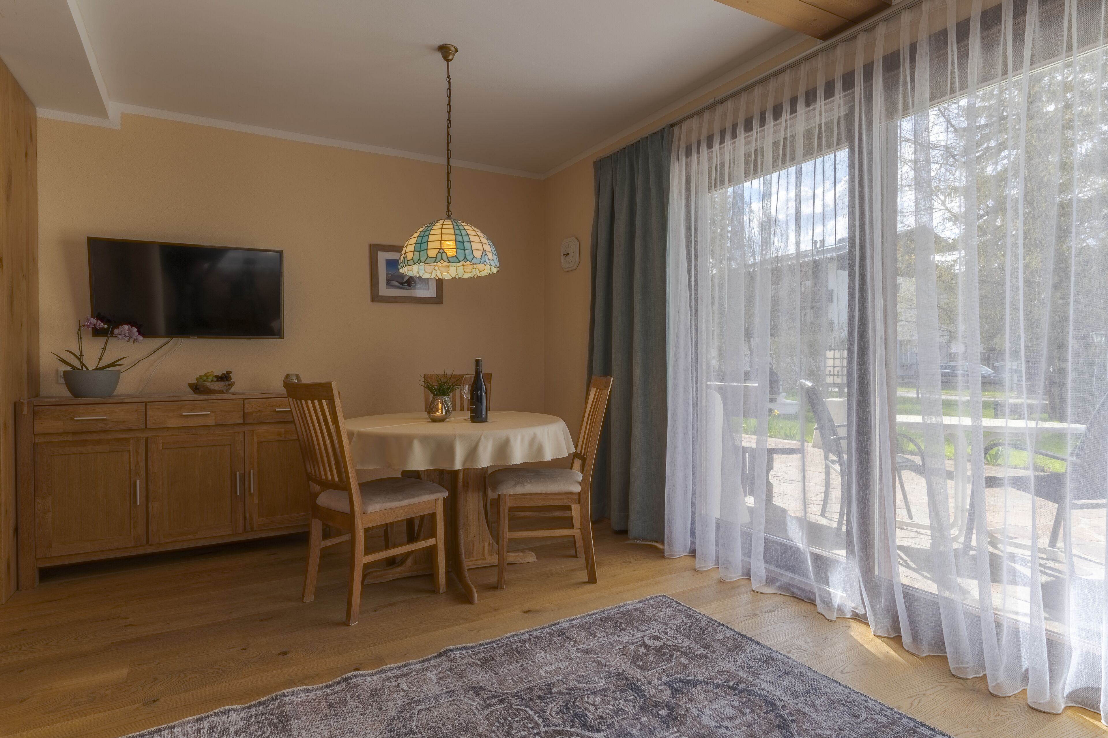 A dining area with a table, chairs, a television, and a window with sheer curtains.