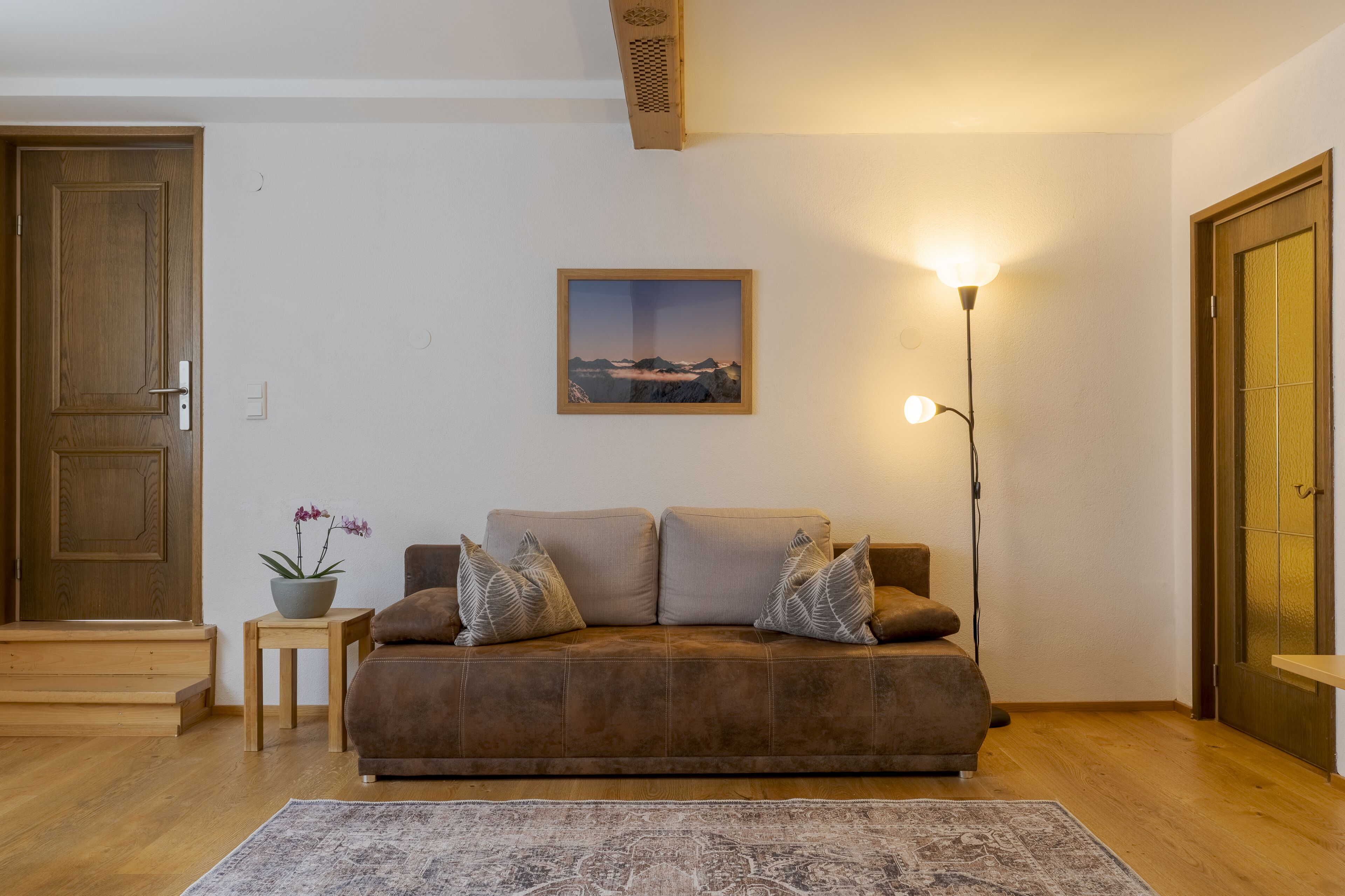 Una sala de estar acogedora con un sofá marrón, una mesita auxiliar con una planta, un cuadro enmarcado en la pared y una lámpara de pie alta.