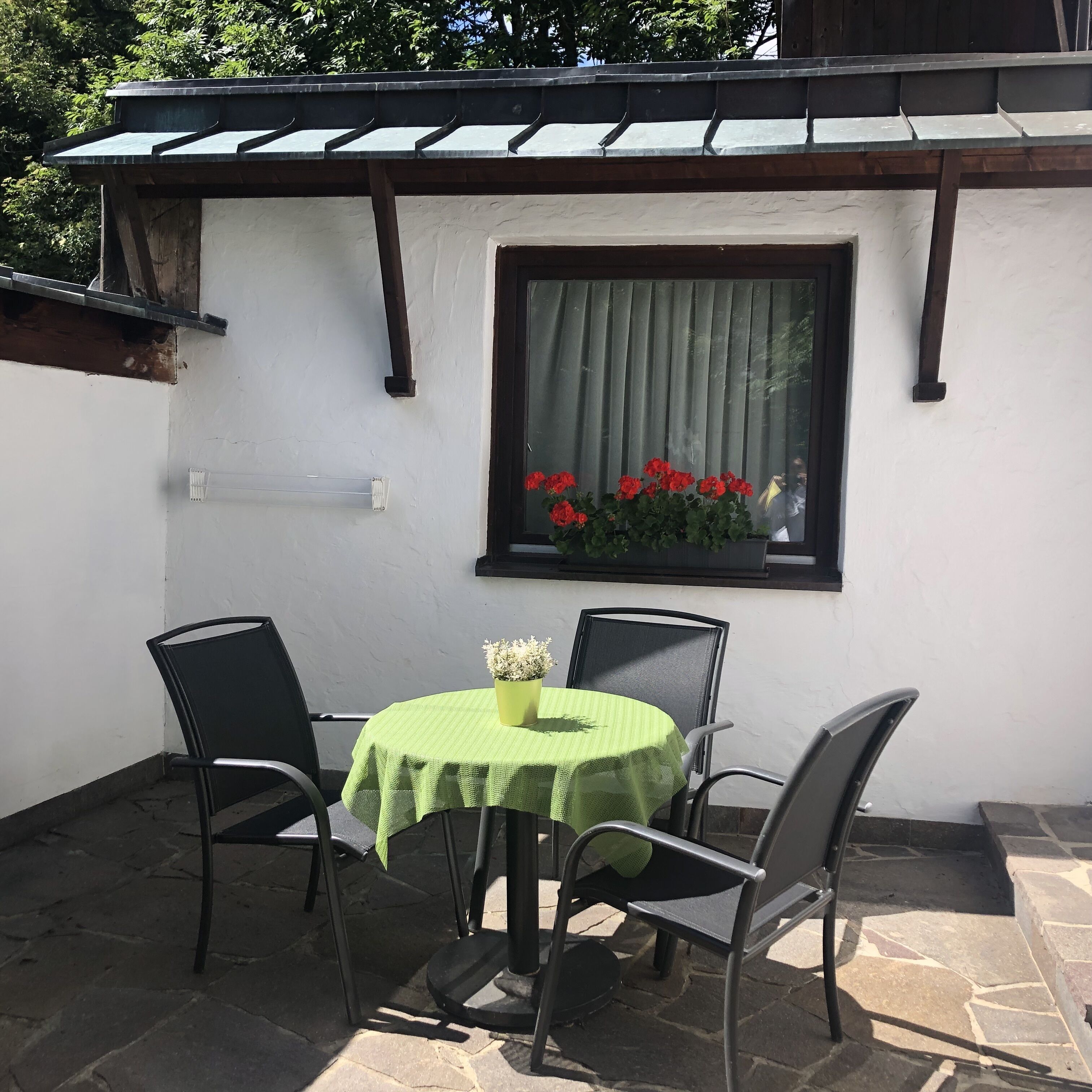 Une petite terrasse extérieure avec une table ronde et quatre chaises, une fenêtre ornée de fleurs rouges et un mur blanc surmonté d’une poutre en bois sombre, au-dessus de la fenêtre.
