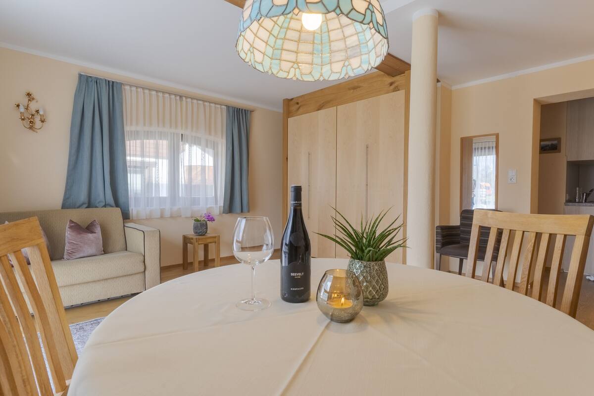 Un comedor con una mesa redonda, una botella de vino y una vela. Se ve un sofá y una pequeña mesa con una planta al fondo.