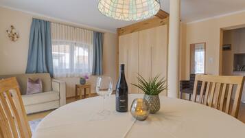 A dining area with a round table, a wine bottle, and a candle. There is a sofa and a small table with a plant in the background.