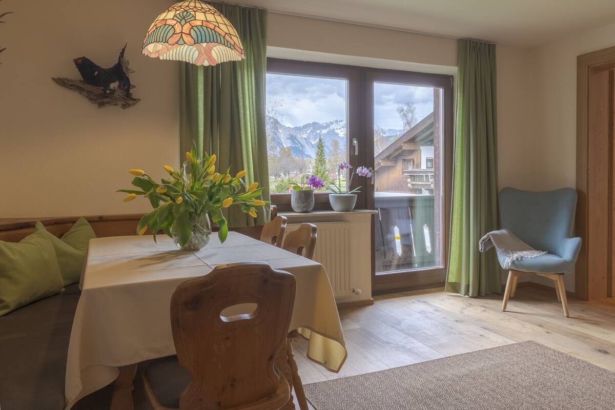 Comedor con una mesa lista para cuatro, una silla, un florero con flores y vistas a montañas a través de un ventanal grande.