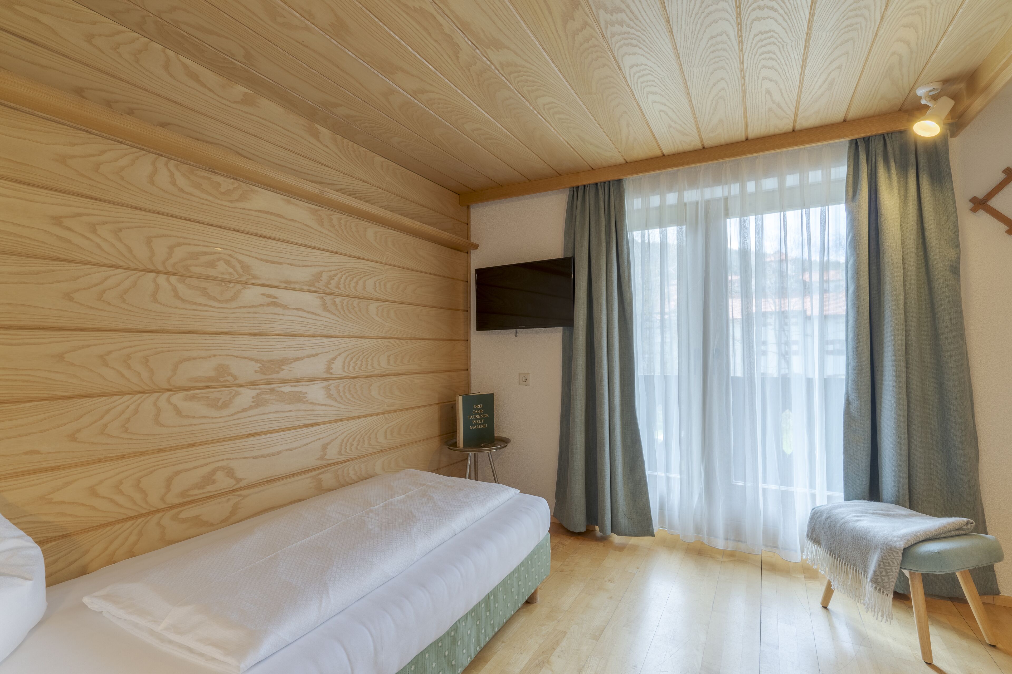 A room with wooden panel walls, a bed, a small table with a TV, and a window with curtains.