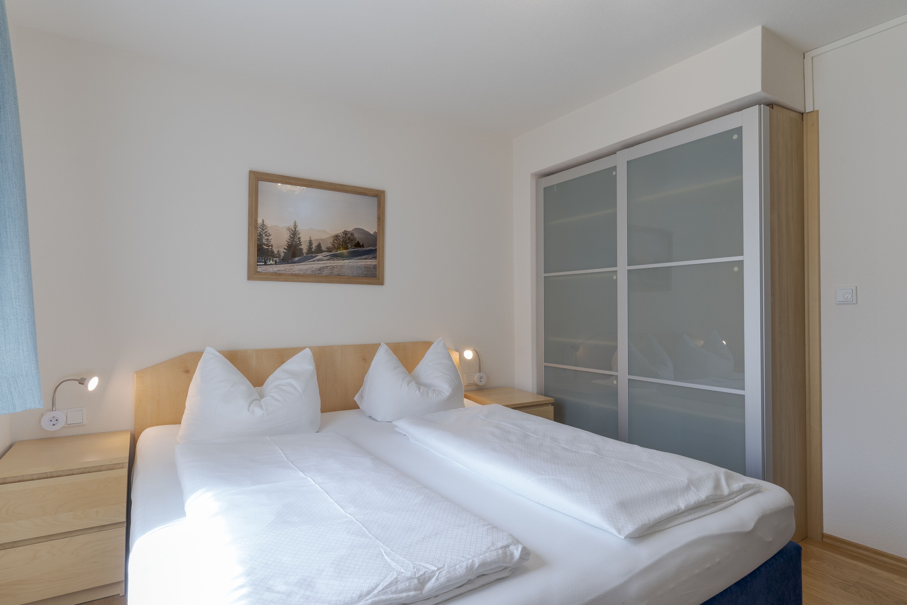Ein ordentlich bezogenes Bett mit weißen Leinen und Kissen, ein Nachttisch mit Lampe und ein gerahmtes Bild an der Wand.