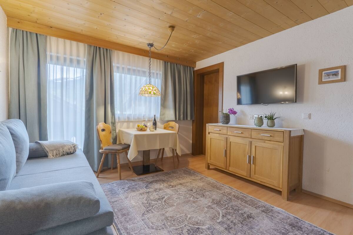 Una sala de estar con un sofá, una mesa de comedor con sillas, un televisor montado en la pared y un mueble de madera.