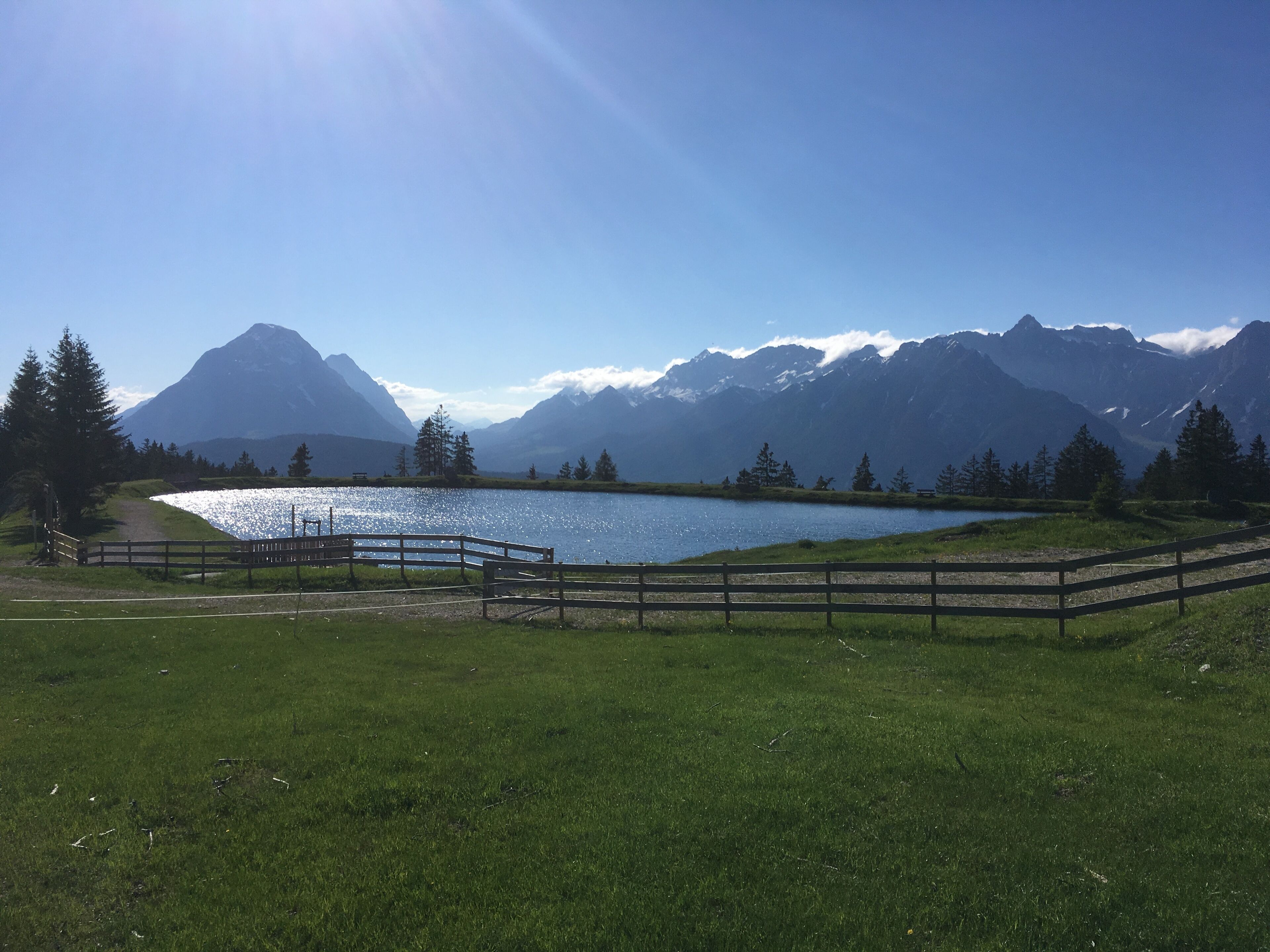 A serene lakeside view with a wooden fence, a clear sky, and mountains in the background.