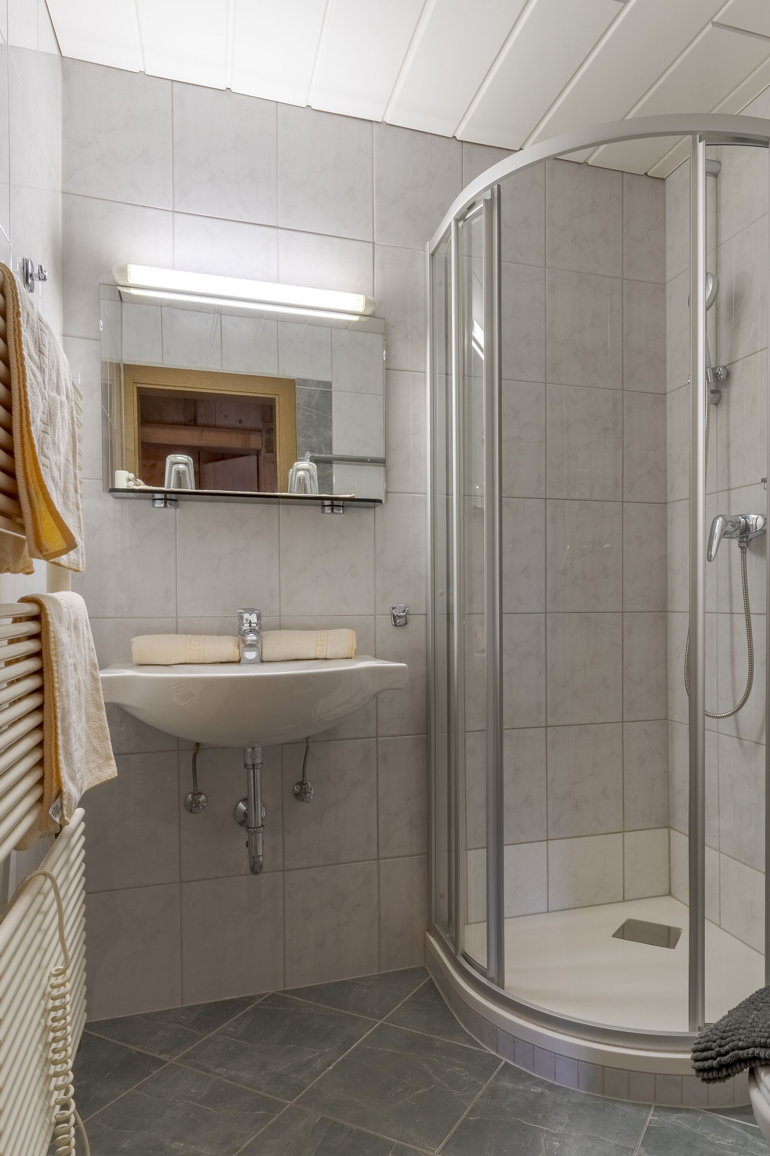 Un baño moderno con ducha envuelta en vidrio, un lavabo blanco con espejo y un perchero con toallas.