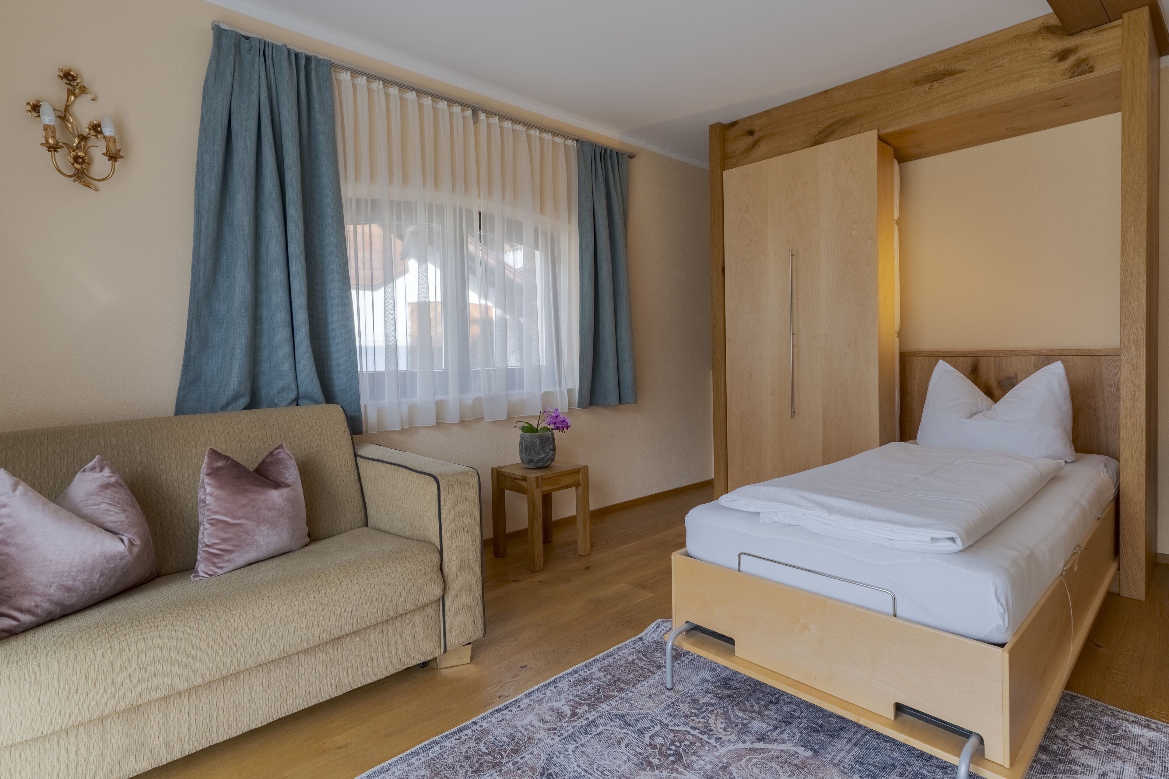 A bedroom with a bed, a sofa, a small table with a vase, and a window with curtains.