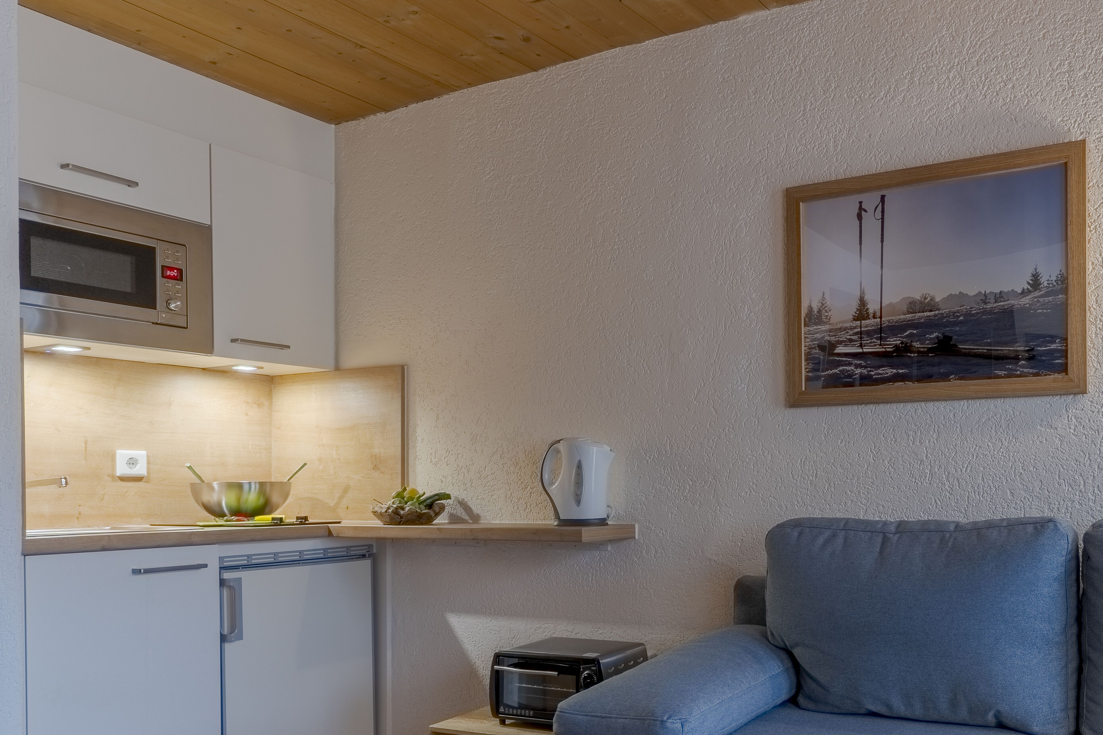 Una cucina compatta con forno a microonde, tostapane e una ciotola sul bancone. Nell'area soggiorno, un divano blu e un quadro appeso al muro.