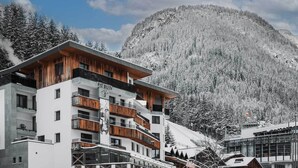 A modern multi-story building with wooden balconies, set against a snowy mountain backdrop. - Hotel Piz Buin (Ischgl)